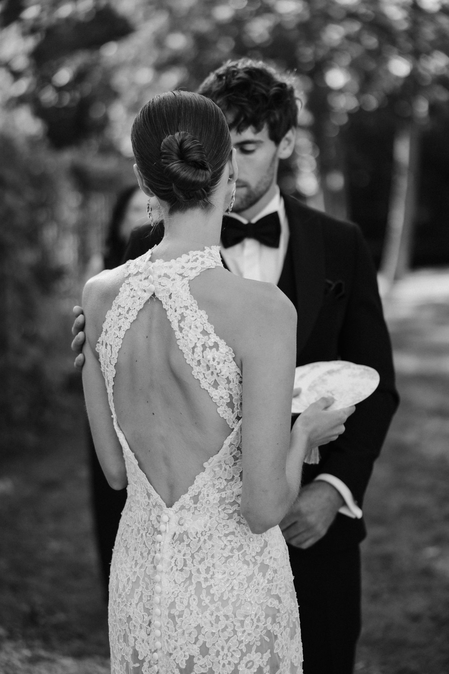 Black and white photo showing the open lace back detail of a bride's gown as groom embraces her.