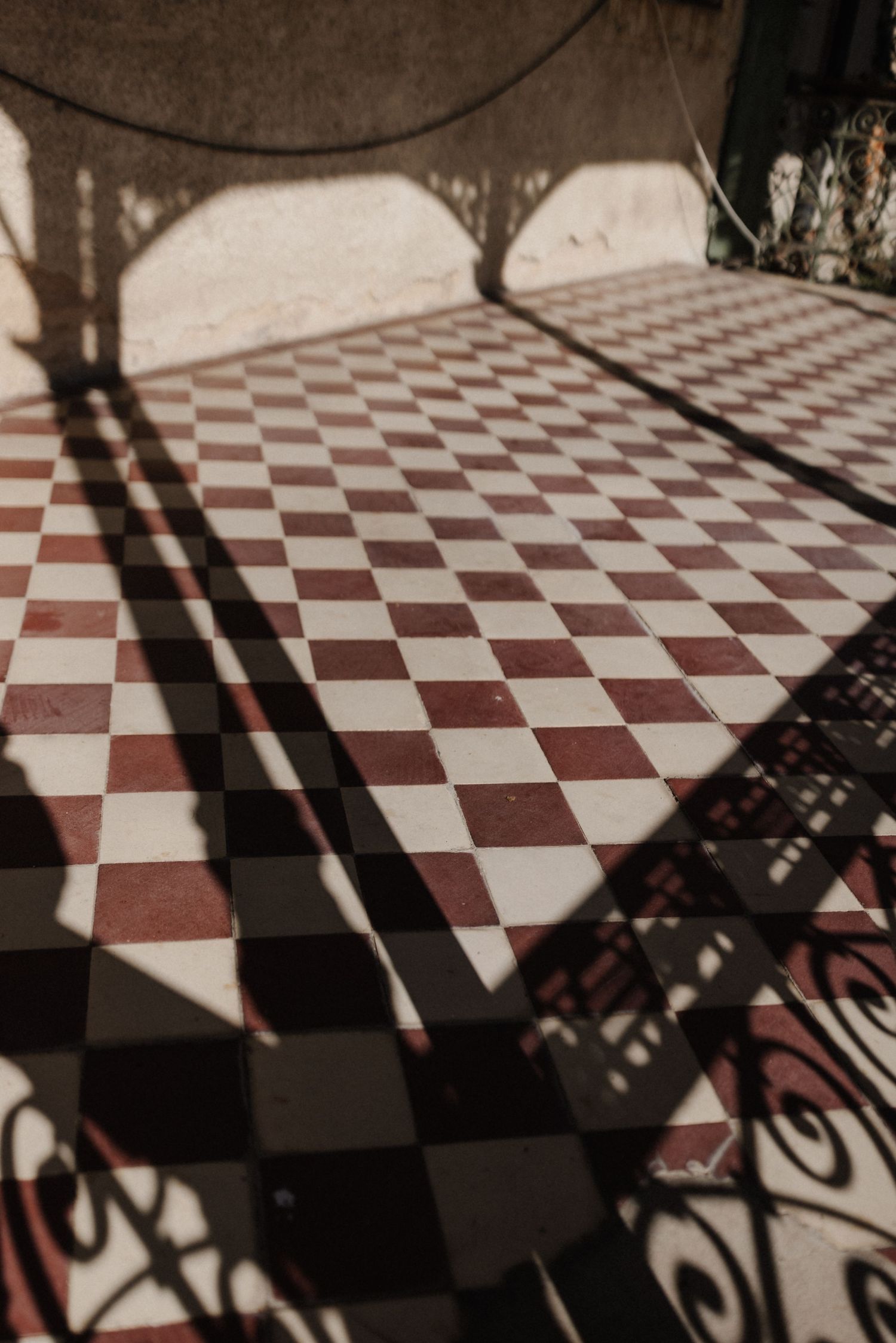 Ornate iron gate shadow cast over red and white checkered tile floor in sunlight.