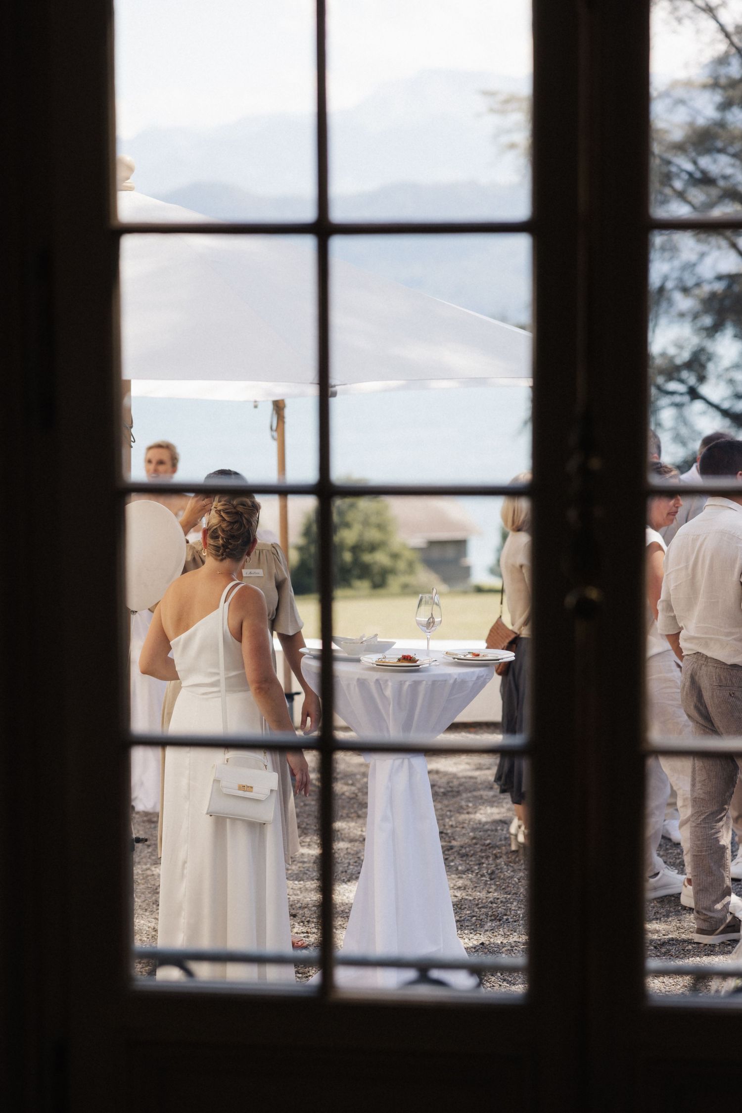 Wedding reception guests socializing outdoors viewed through a classic French door window frame.