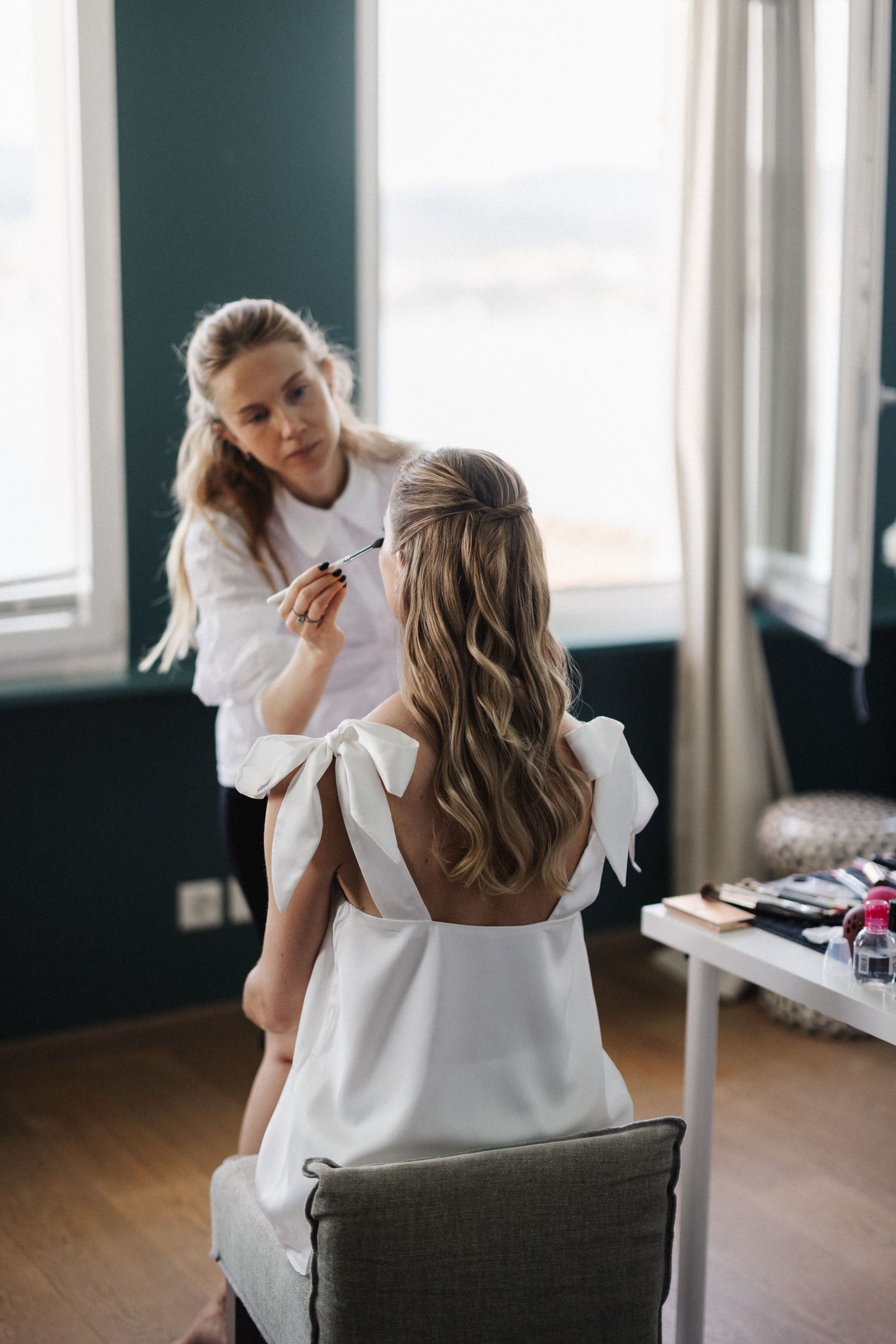 Makeup artist applying cosmetics to a bride in a white dress seated by a bright window.