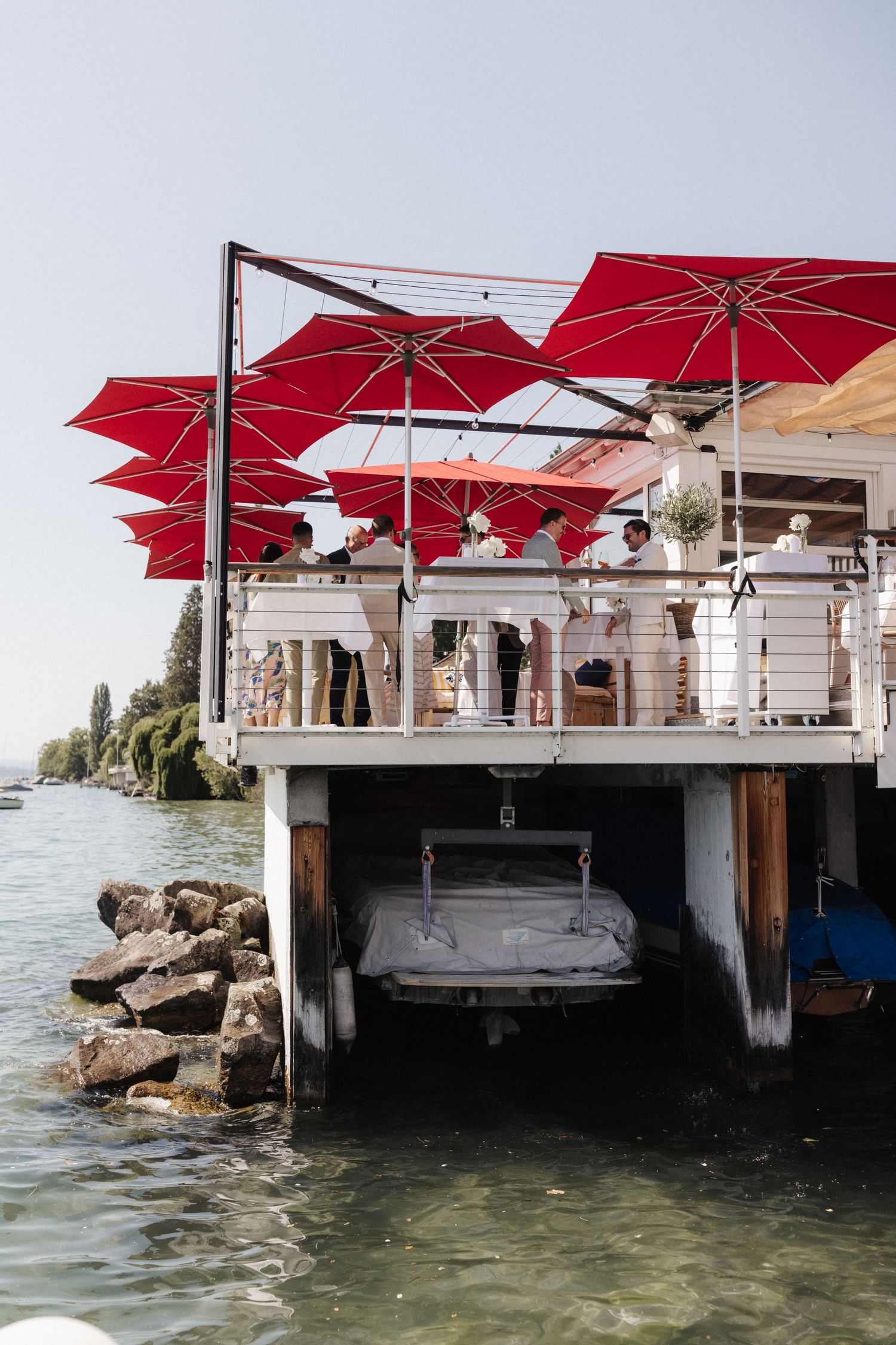 Waterfront venue with red umbrellas, white railings, and a boat stored beneath the deck.