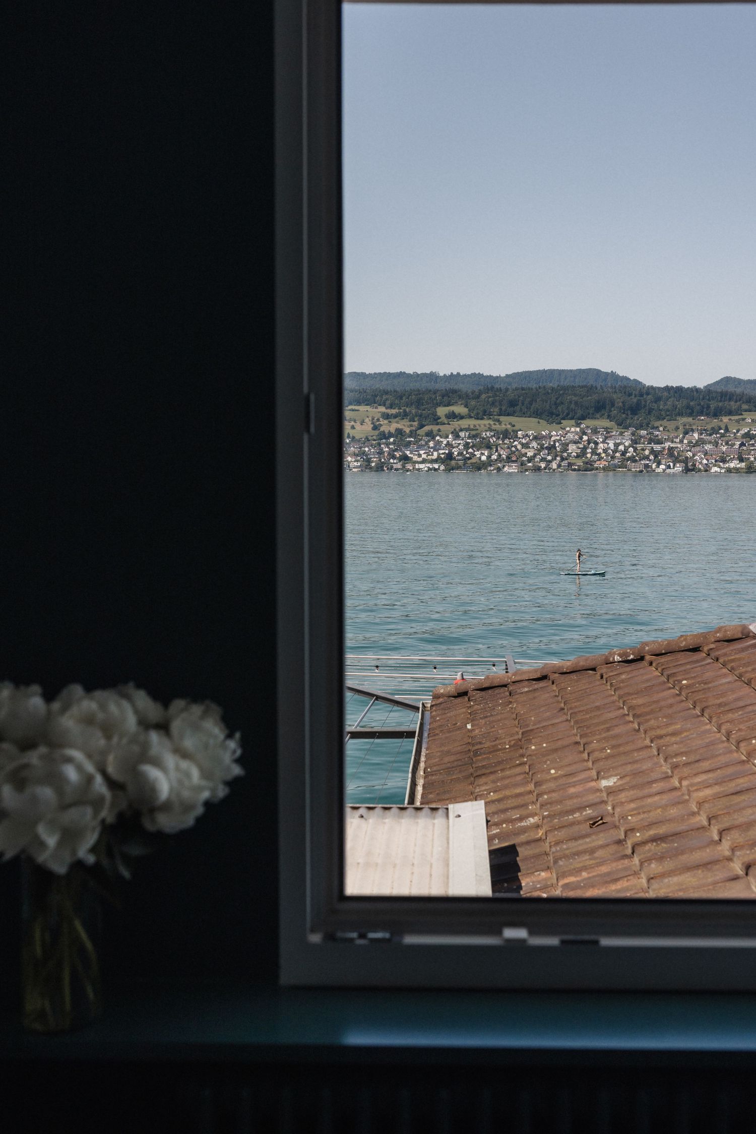 View through a dark window of a lake with a paddleboarder and white peonies in foreground.