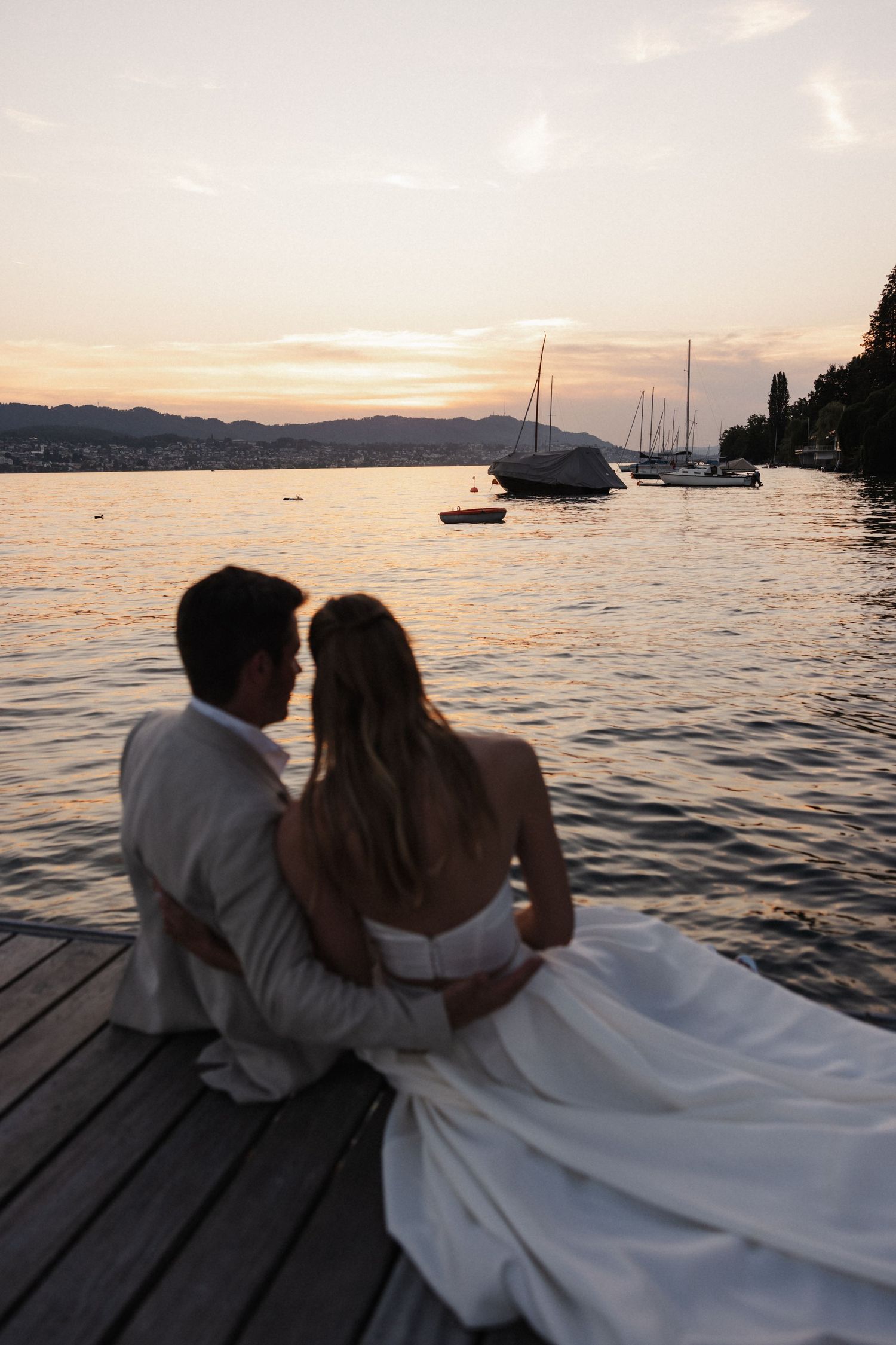 Bride and groom sit embracing on a wooden dock at sunset, overlooking a calm lake with sailboats.