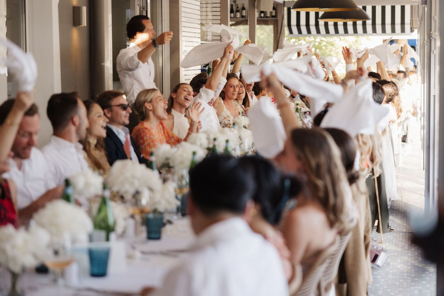Wedding guests raise white napkins in celebration at a long reception table adorned with white floral centerpieces.