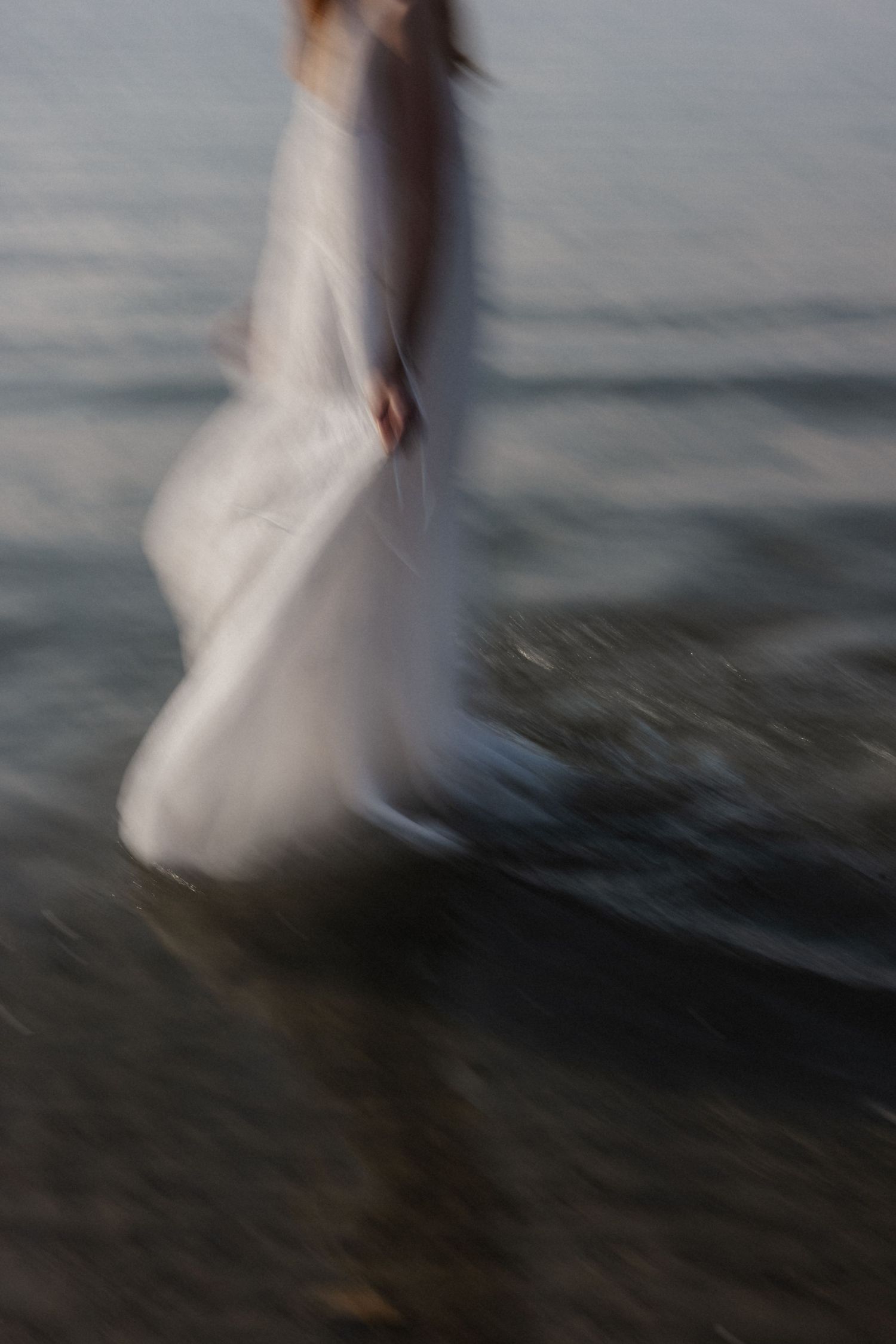Blurred motion shot of woman in white dress walking through ocean waves at dusk.