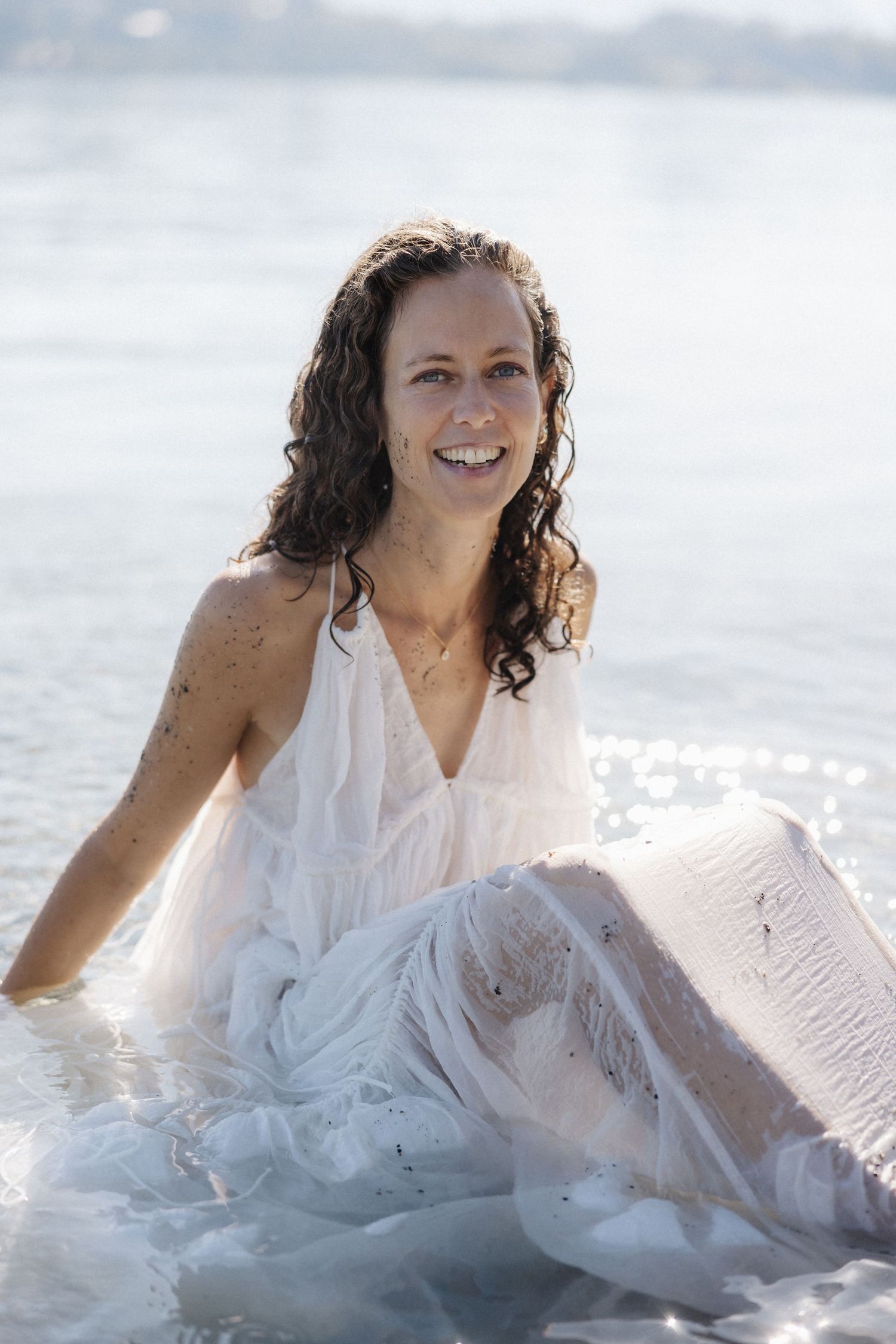 Smiling woman in wet white dress sitting in shallow water, looking at camera happily.