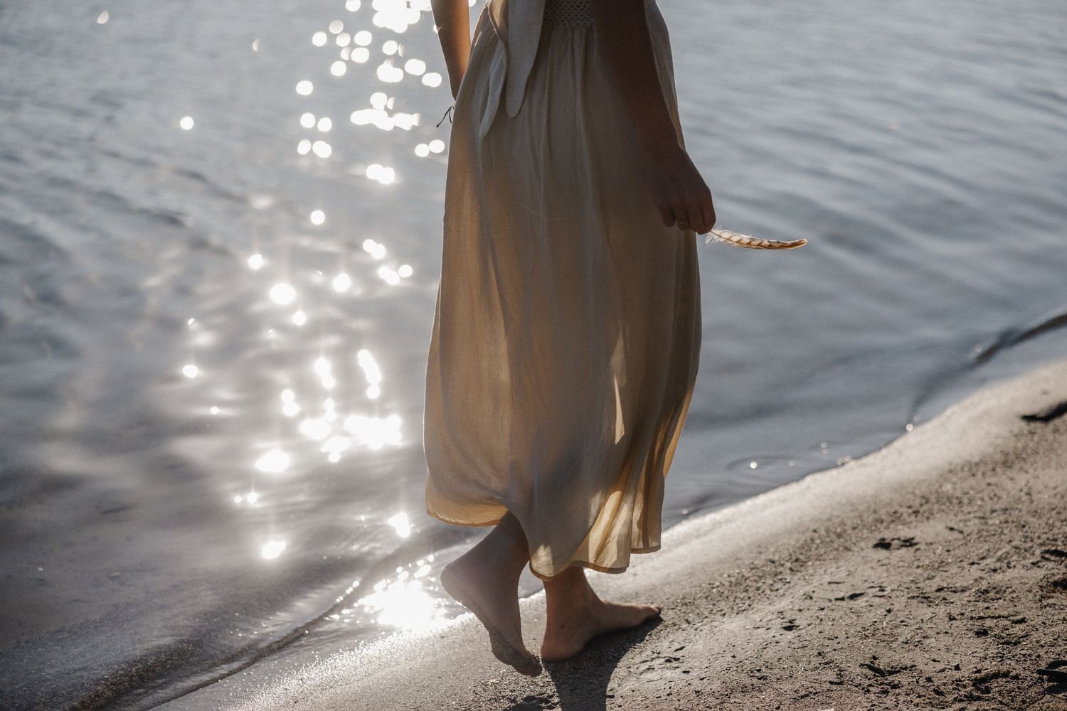 Barefoot woman in flowing beige dress walks along sunlit shimmering shoreline with sparkling water reflections.
