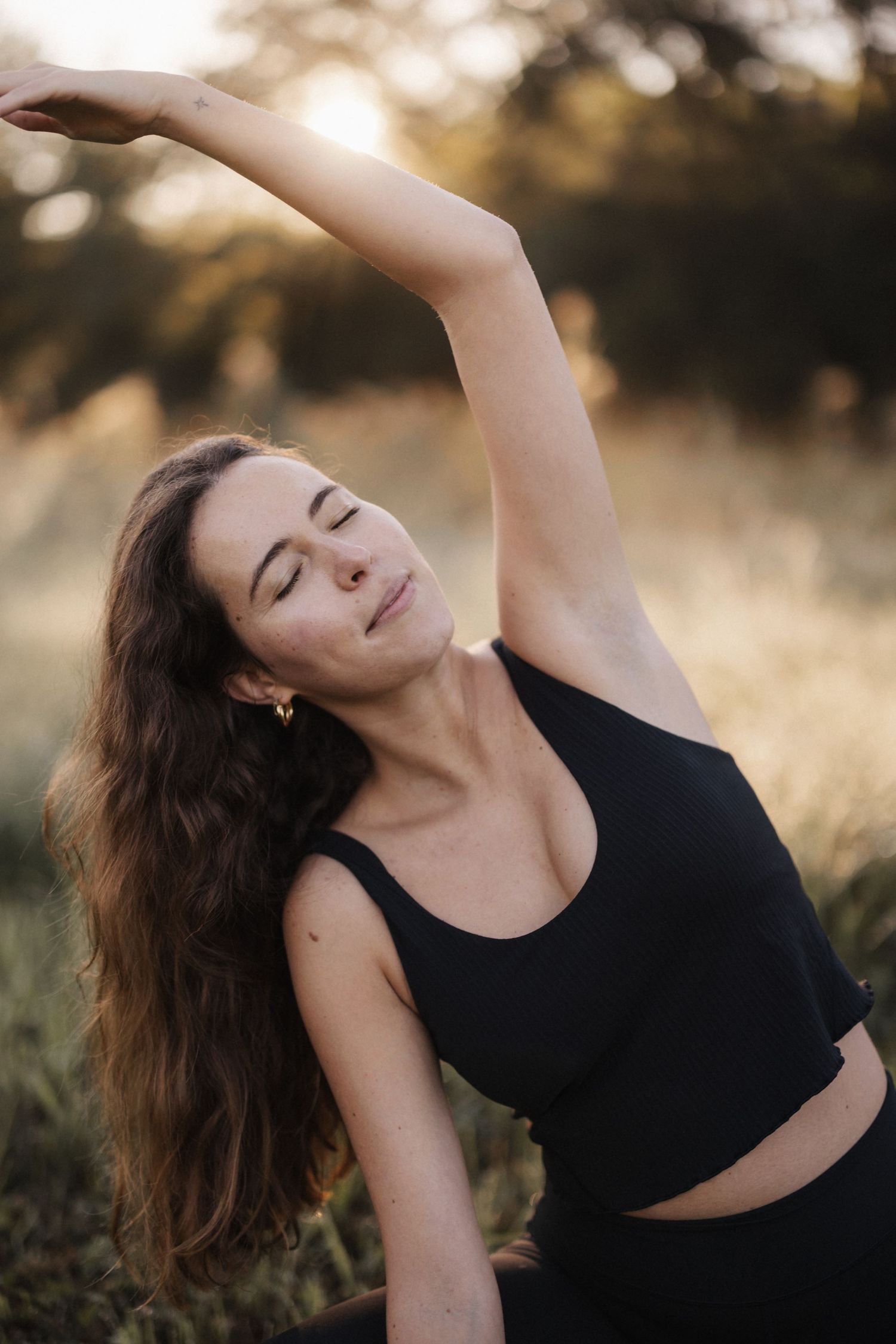 Young woman in black activewear doing a side stretch yoga pose outdoors in a golden sunset field.