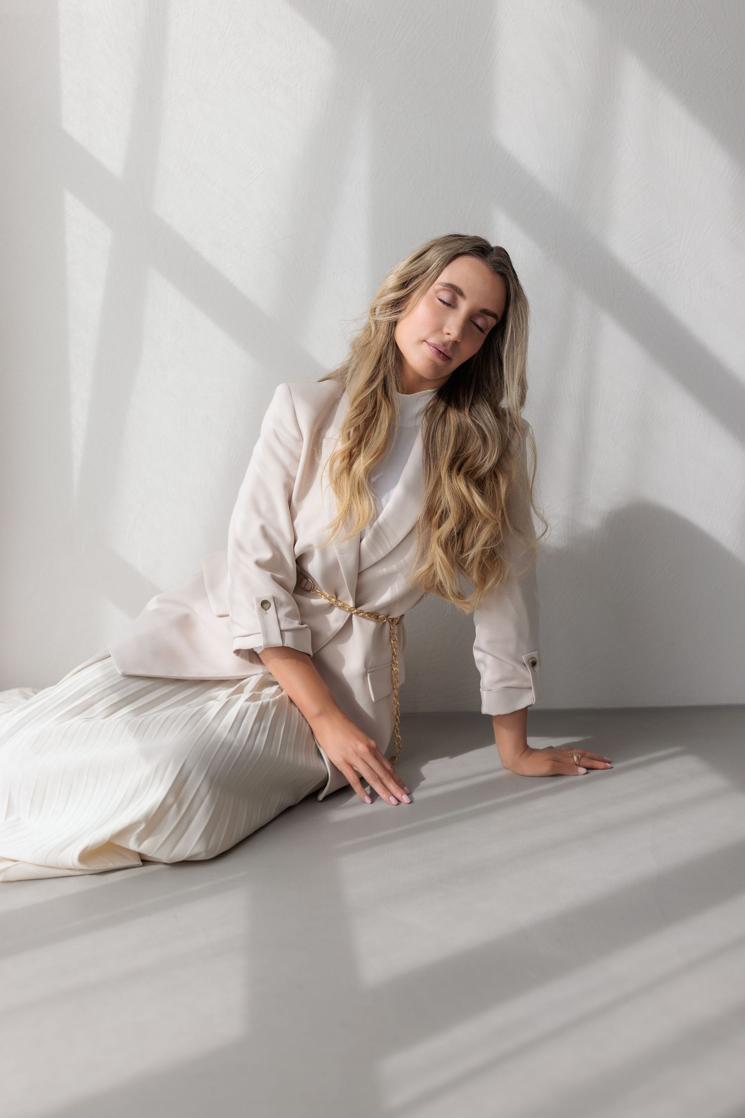 Elegant blonde woman in cream pleated skirt and jacket posing on floor in bright minimalist studio.