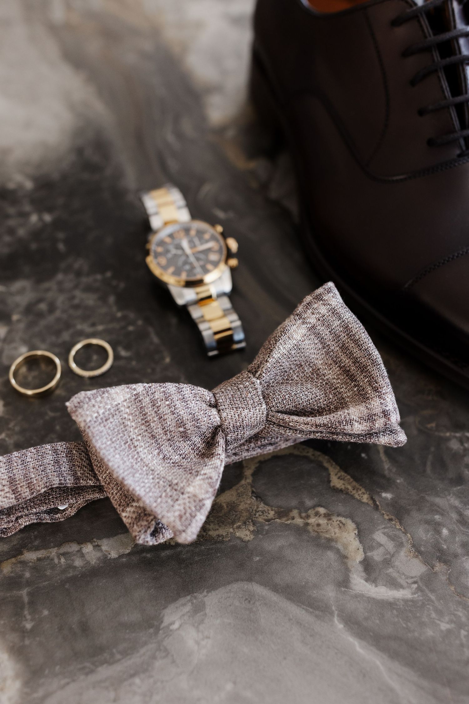 Close-up of plaid gray bow tie, gold wedding rings, luxury watch, and brown dress shoe on marble.