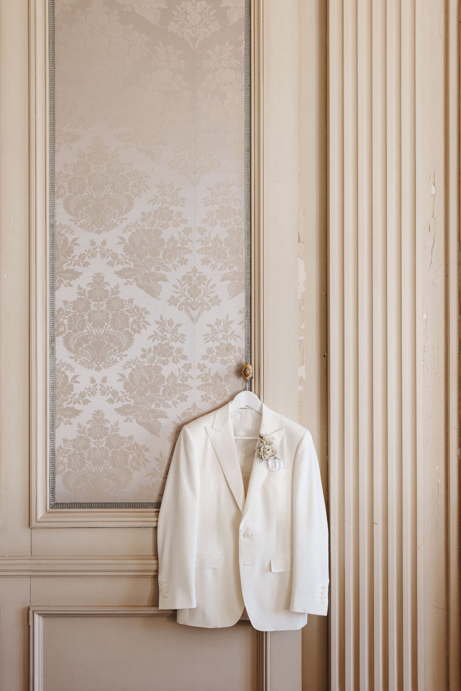 White wedding suit jacket with boutonniere on hanger against elegant beige damask paneled wall.