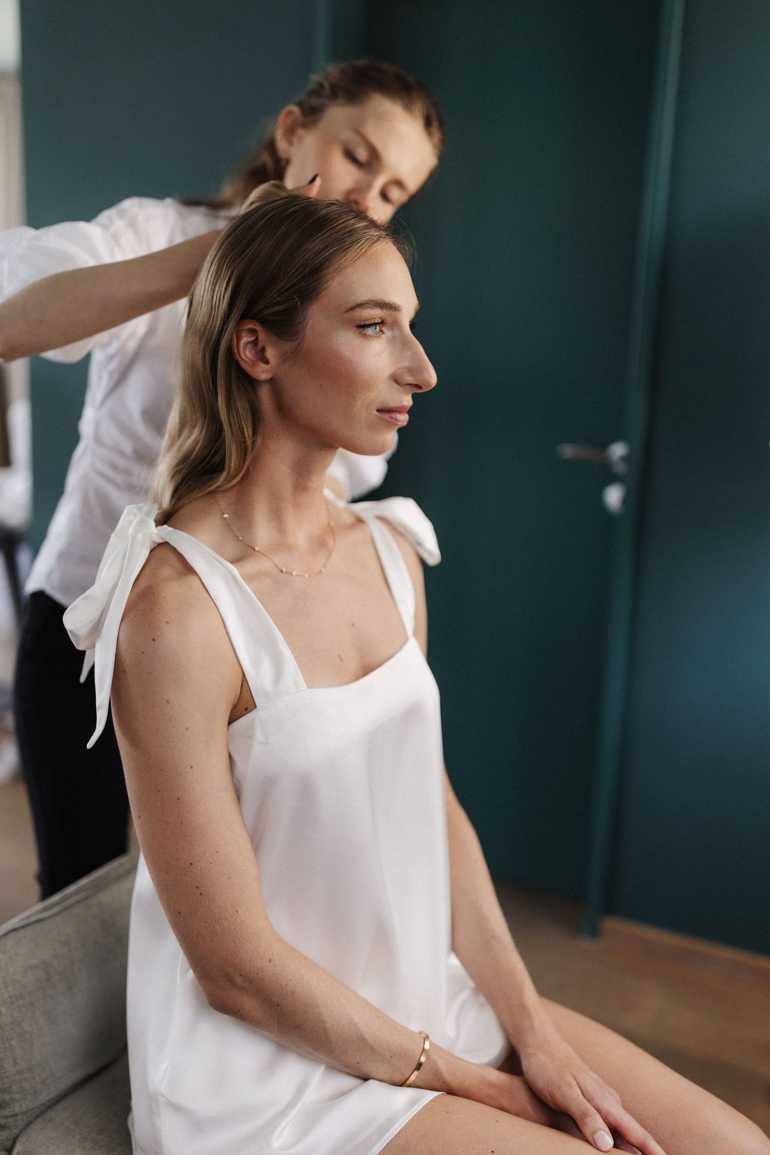 Hairstylist arranging blonde woman's hair in white dress in teal interior.