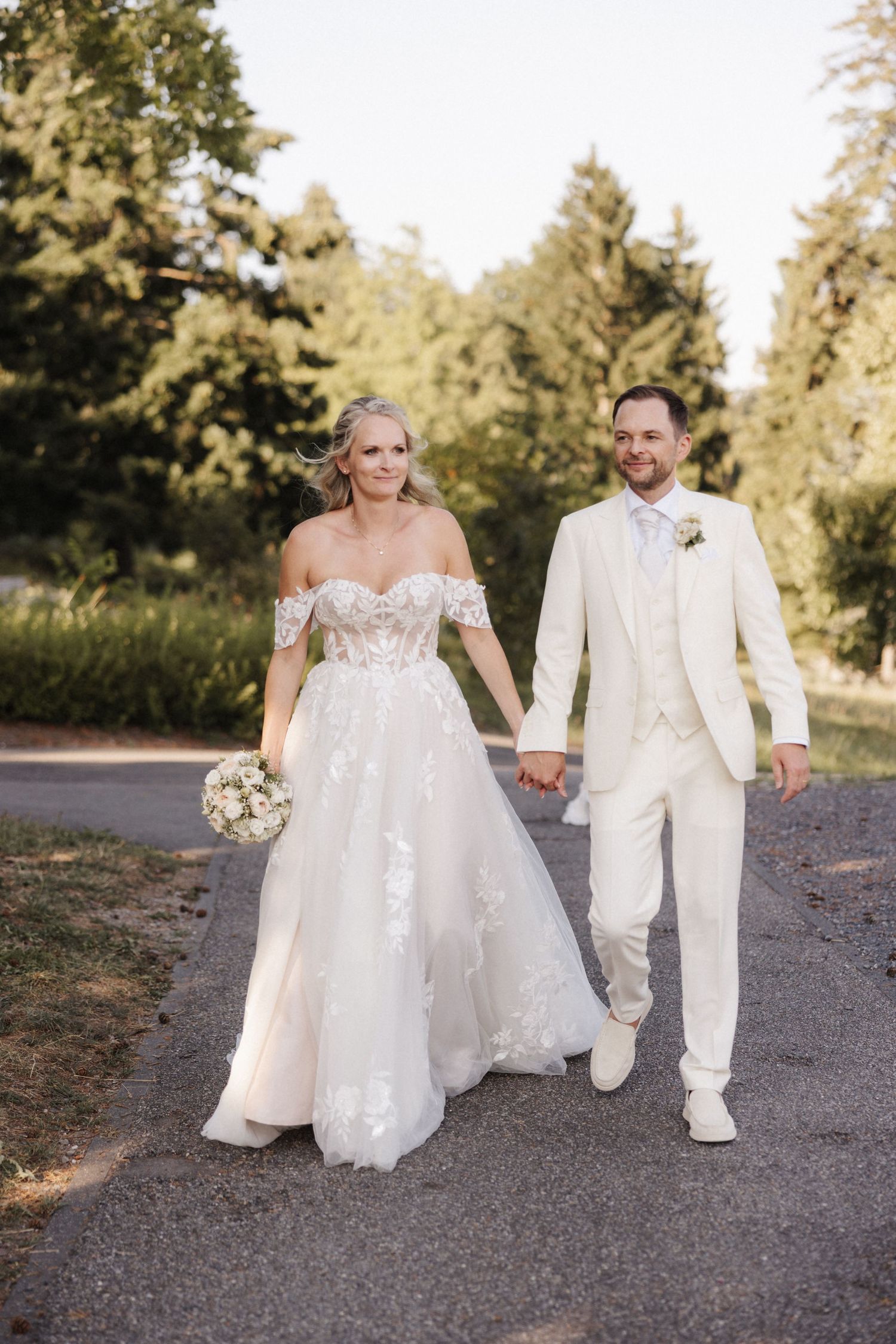 A smiling bride and groom in white attire walk hand-in-hand along a tree-lined path on their wedding day.