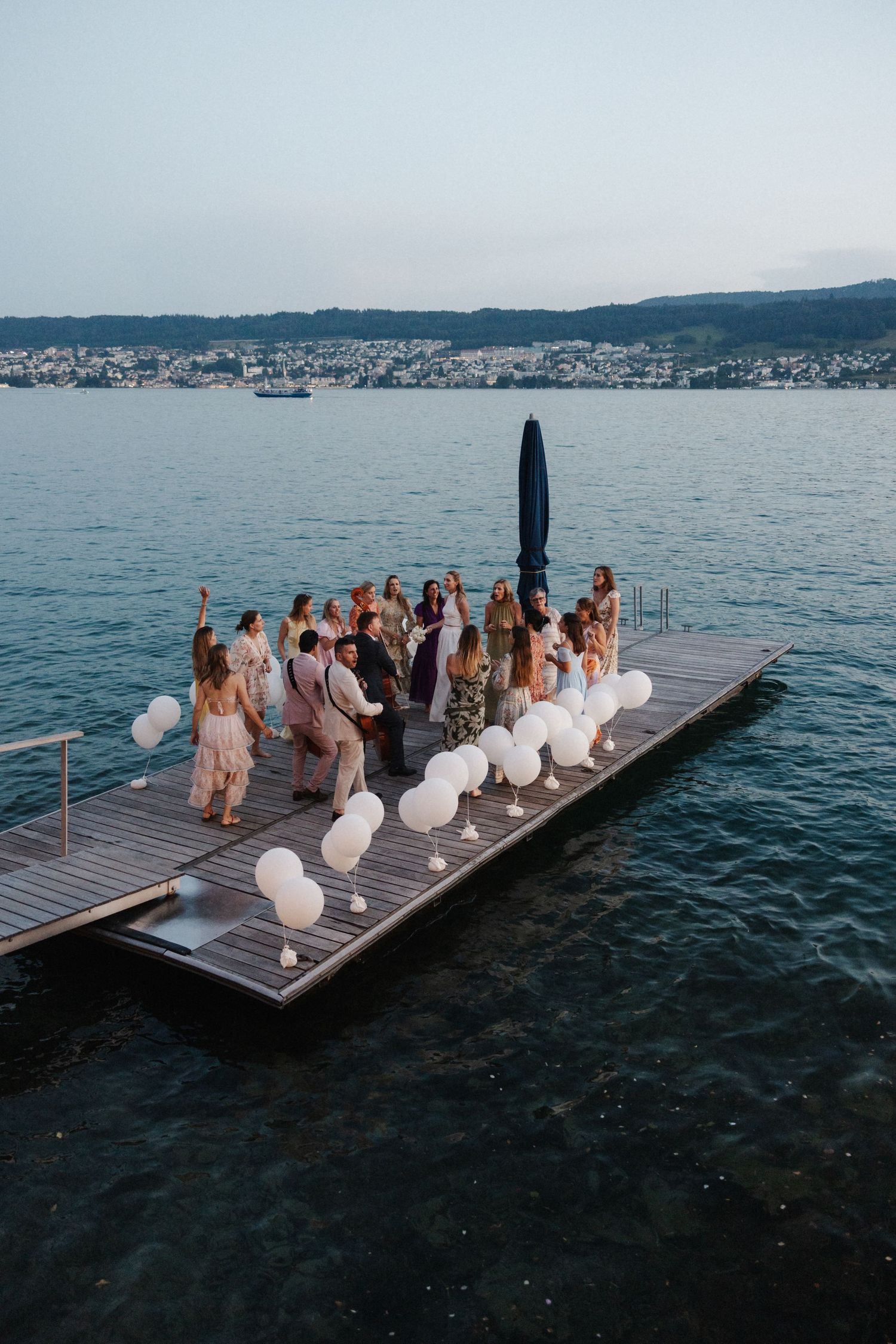 Wedding guests celebrating on a wooden dock over water with white balloons at dusk.
