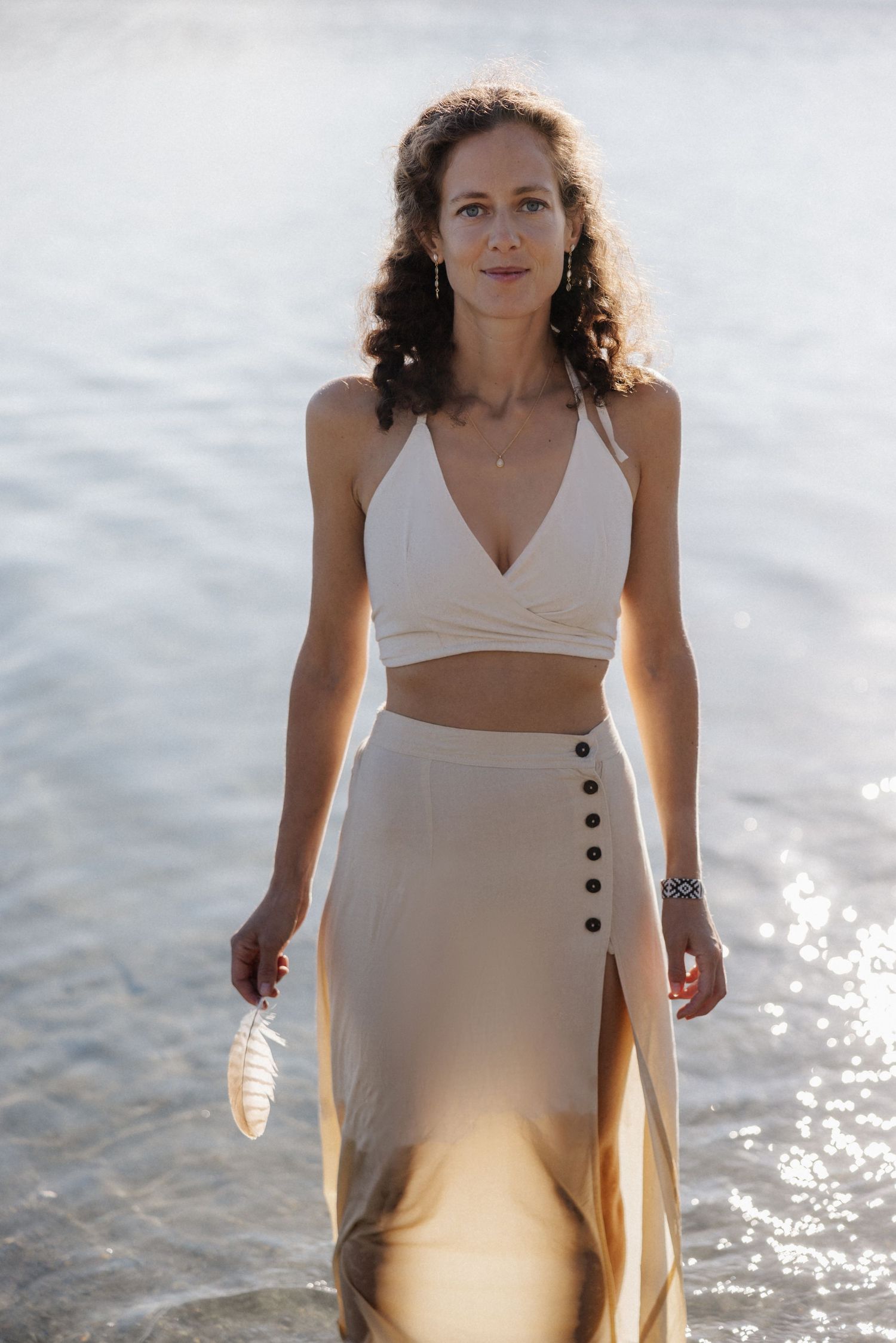 Confident woman in cream crop top and button skirt walks through sparkling shallow water holding a feather.