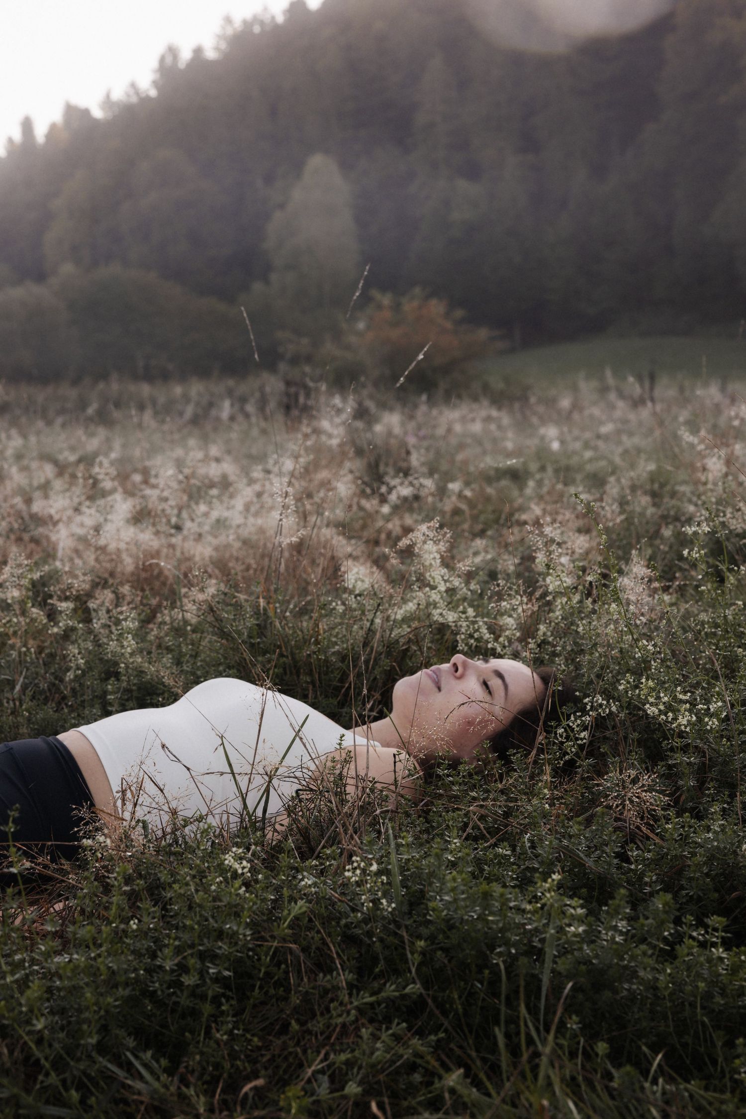 Young woman lying peacefully in a wildflower meadow with misty forested hills in the background.