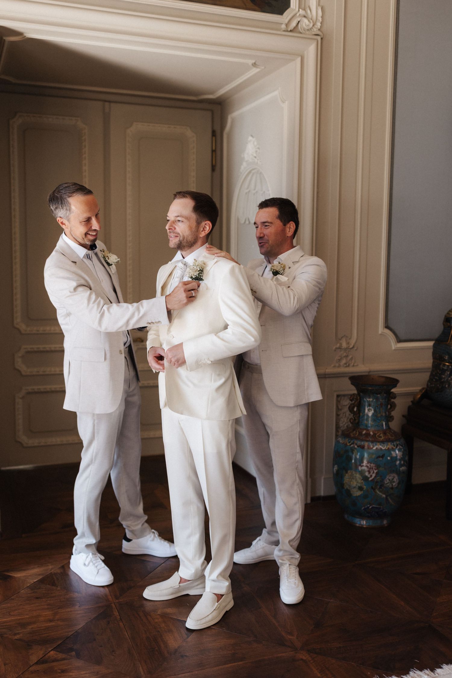 Three men in cream suits helping groom with boutonniere in ornate wedding venue interior.