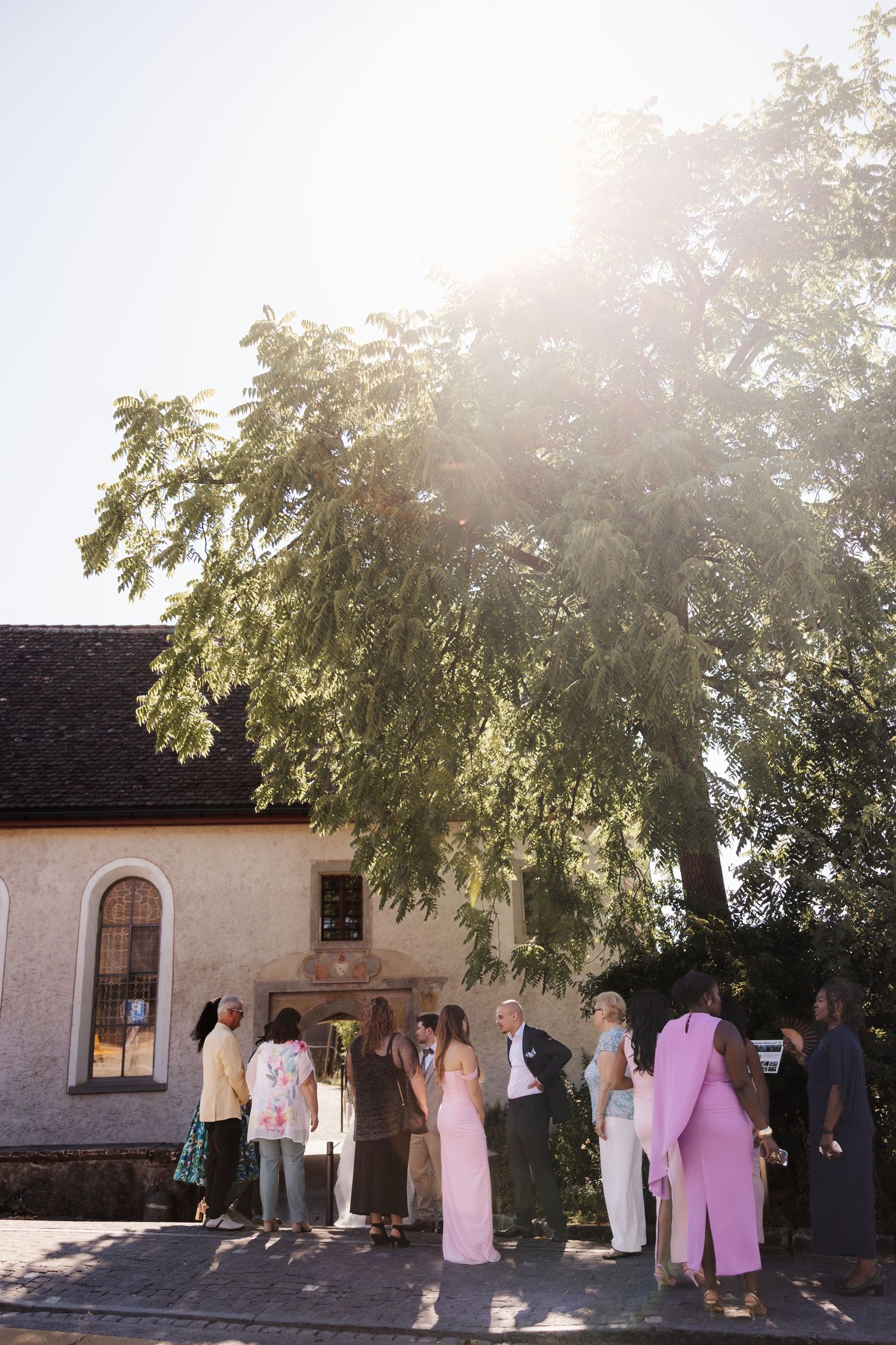 Wedding guests gather outside small stone church under large tree with bright sunlight streaming through branches.