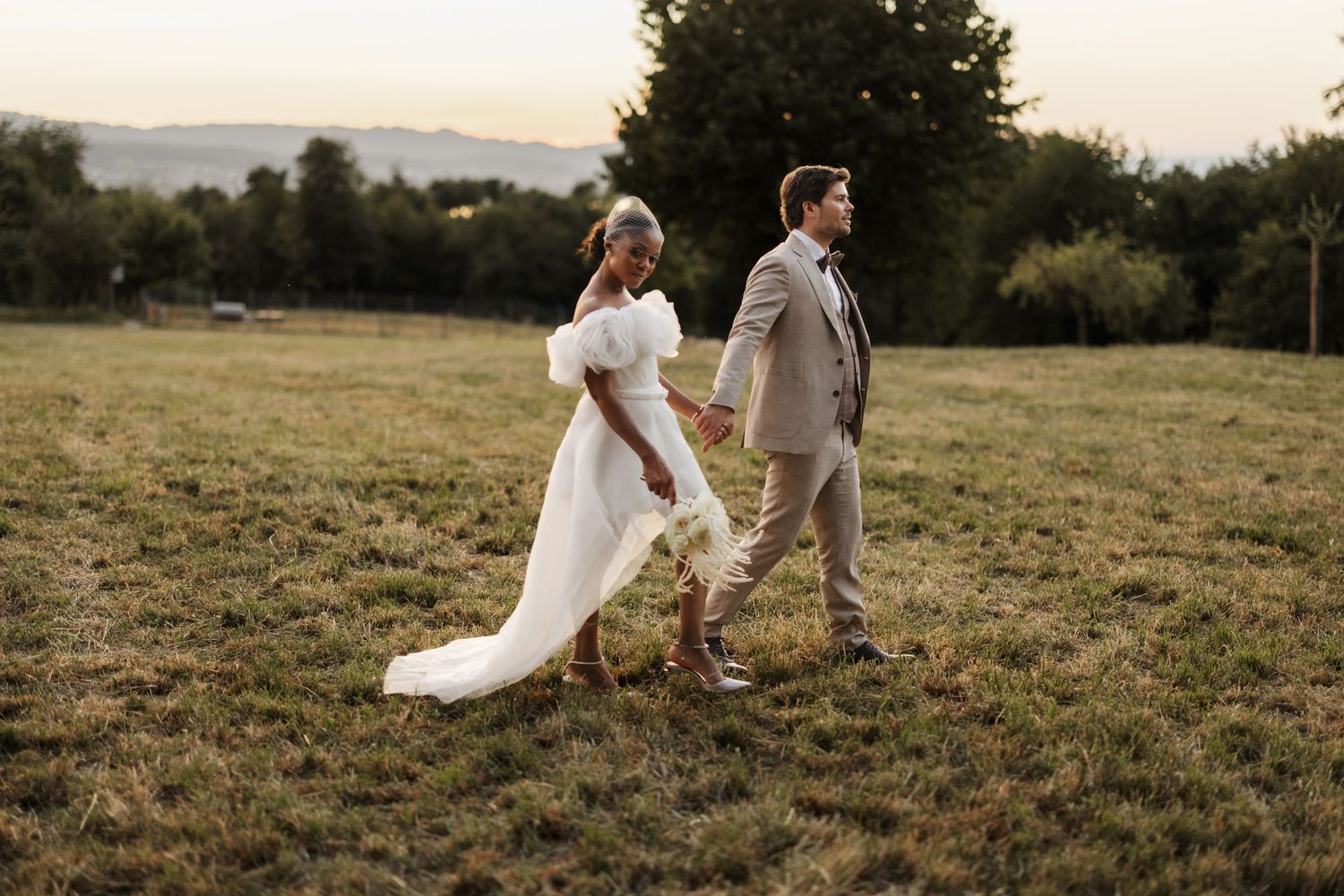 Bride and groom walking hand in hand across a grassy field at sunset, bride in white gown with train.
