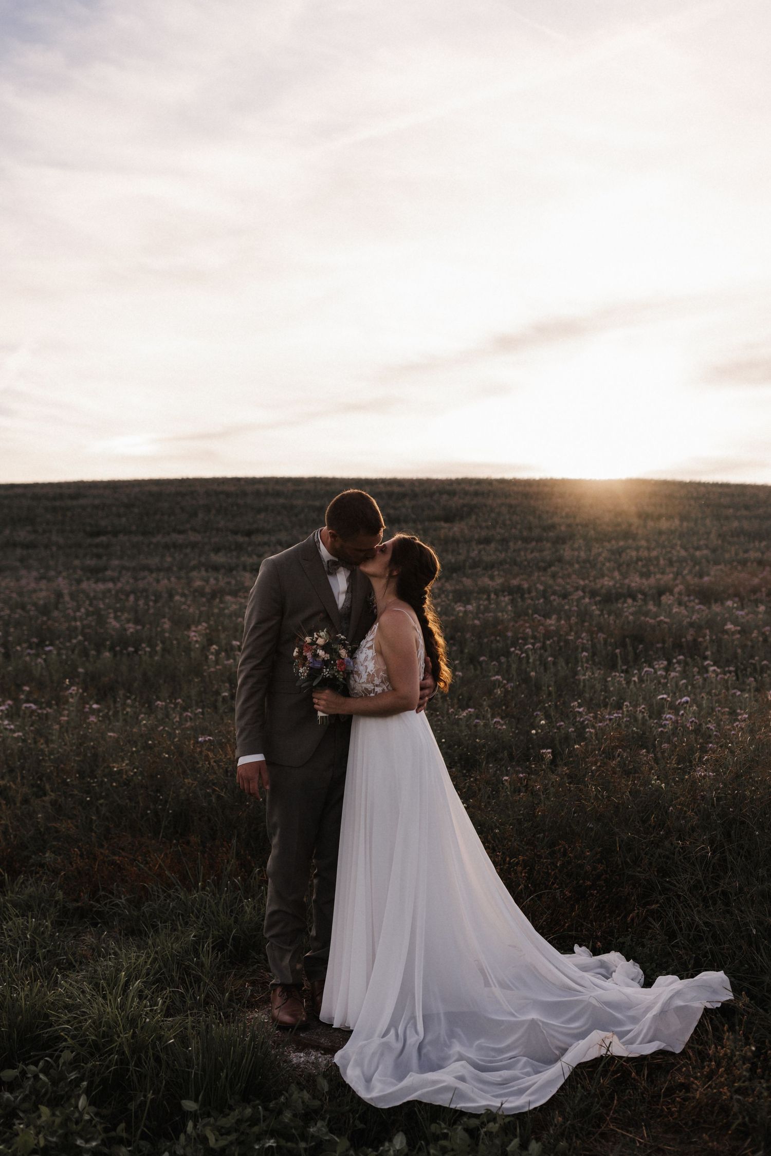 Bride and groom share a romantic kiss in a field at golden hour sunset, bride in flowing white gown.