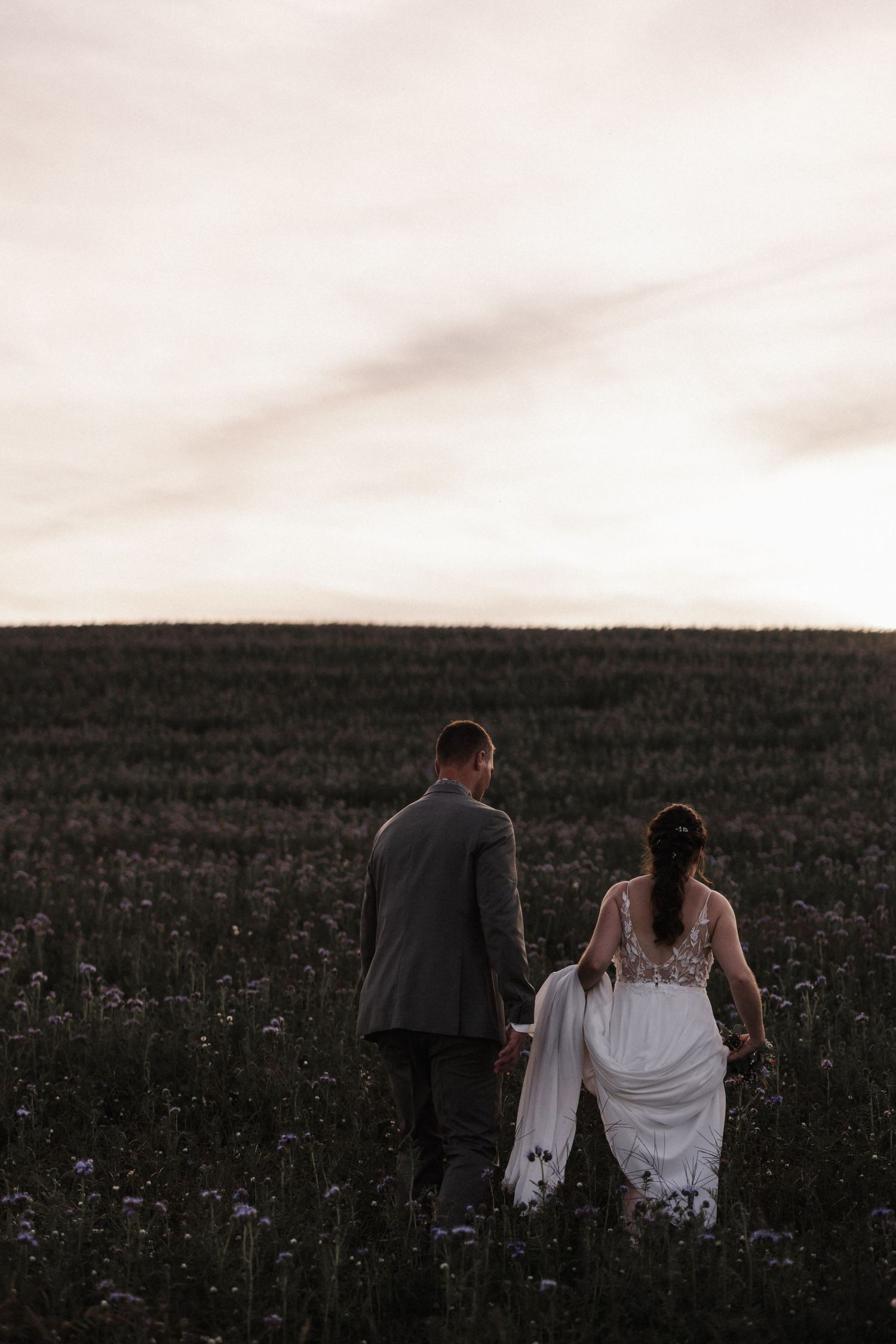 Bride lifts white gown while walking with groom through a vast wildflower field at sunset.