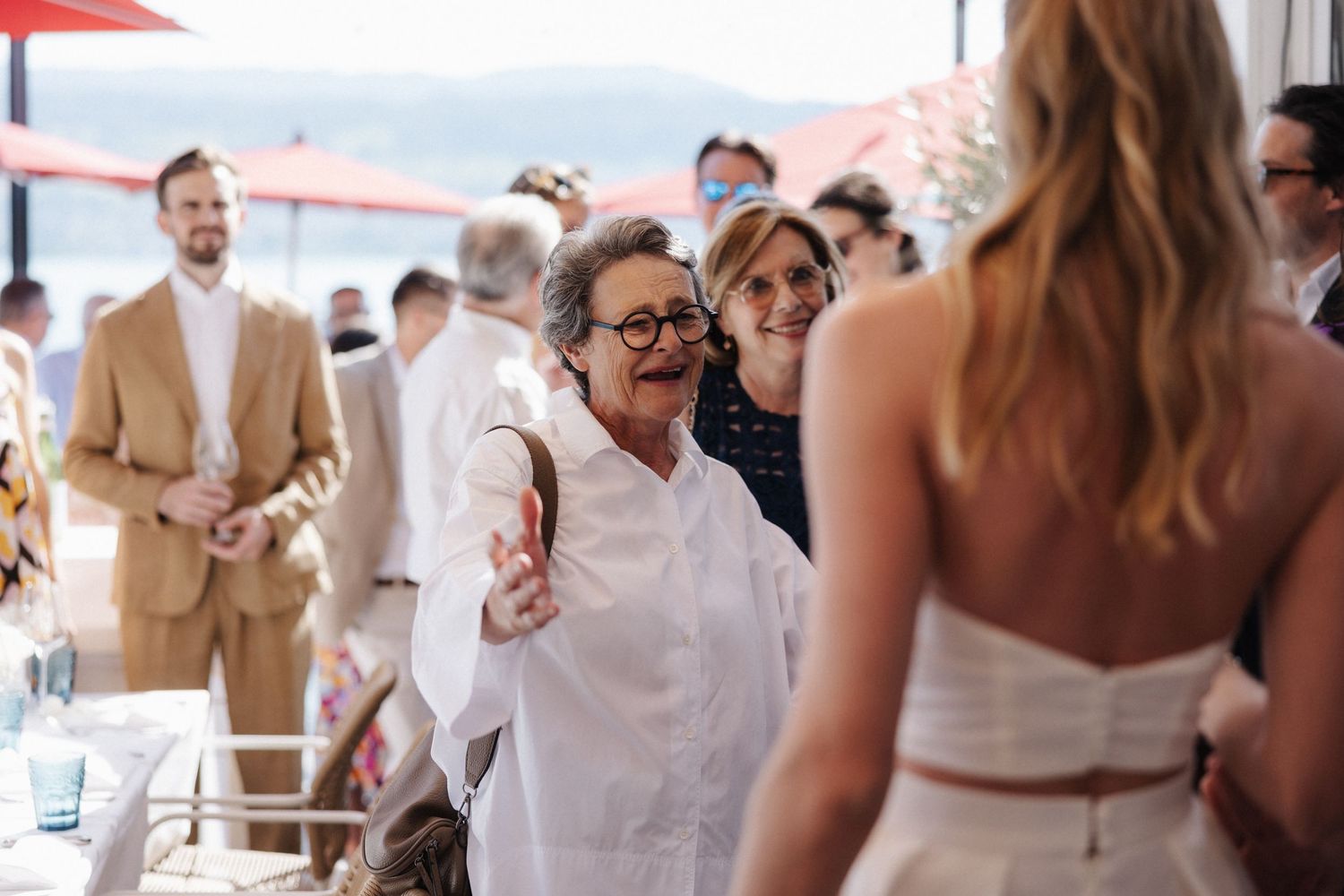 Elderly woman reaching out to embrace the bride at an outdoor lakeside wedding reception.