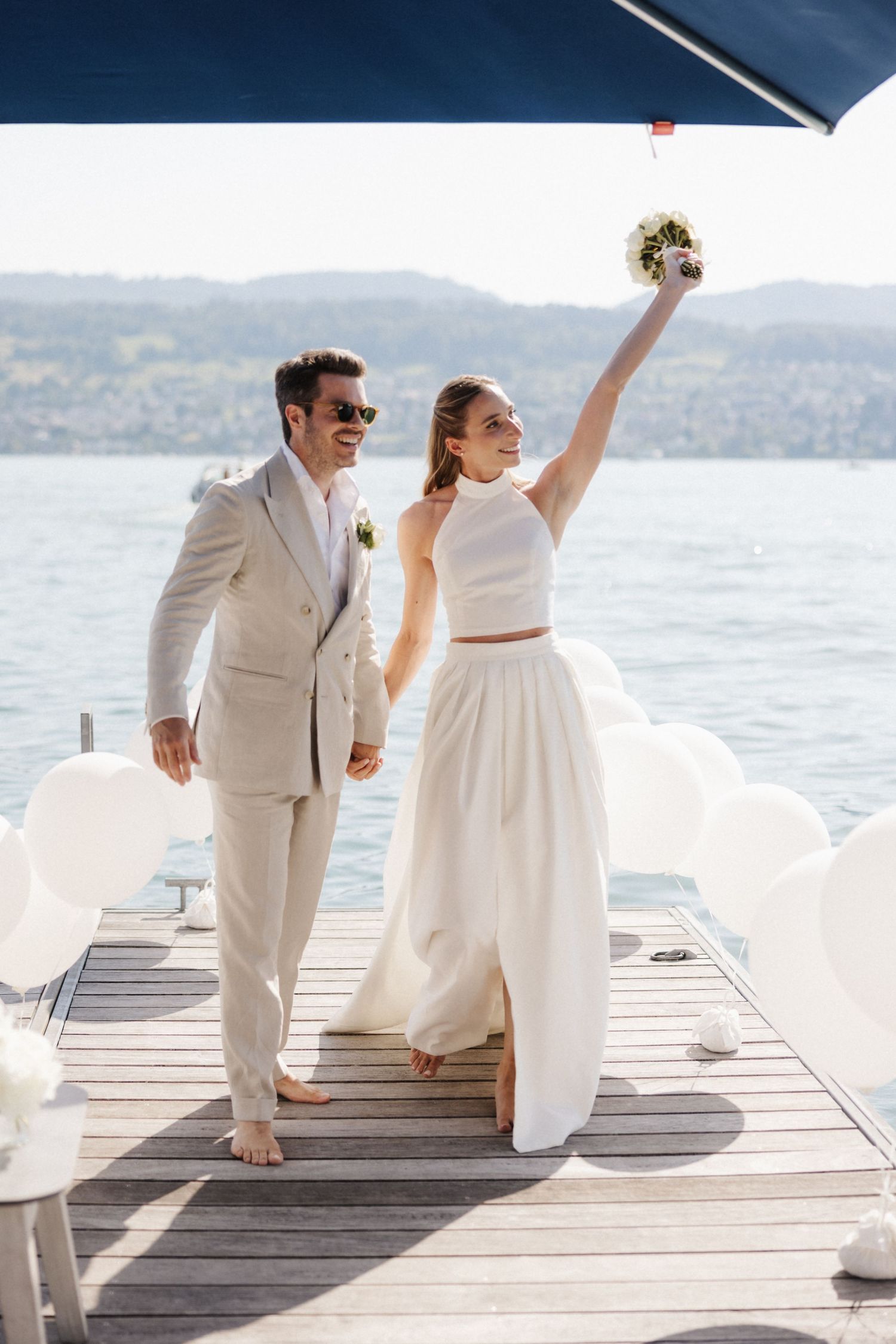Joyful bride raising bouquet with groom on a lakeside dock with white balloon decorations.