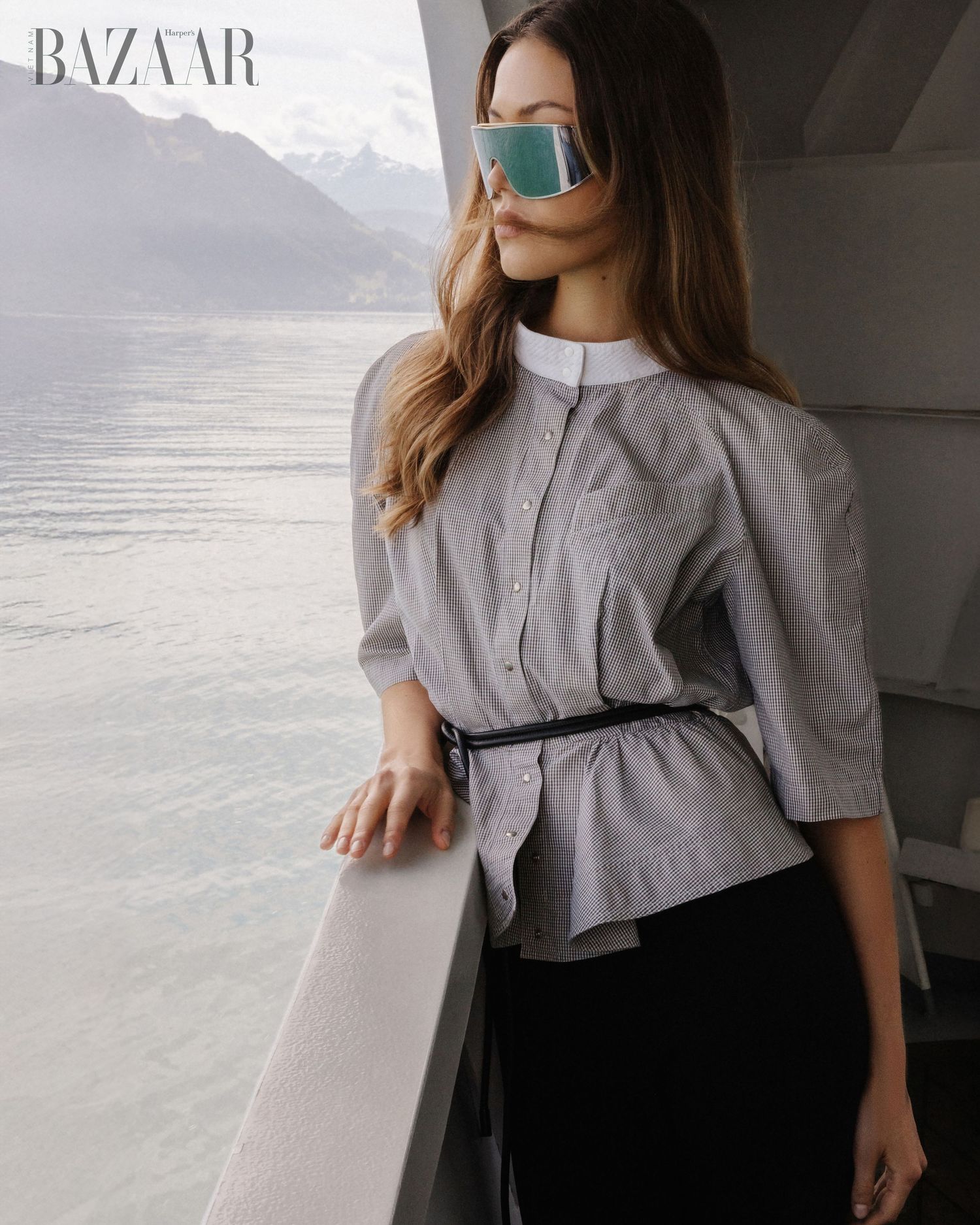 Woman in grey belted peplum blouse and mirrored sunglasses stands on a scenic boat deck.