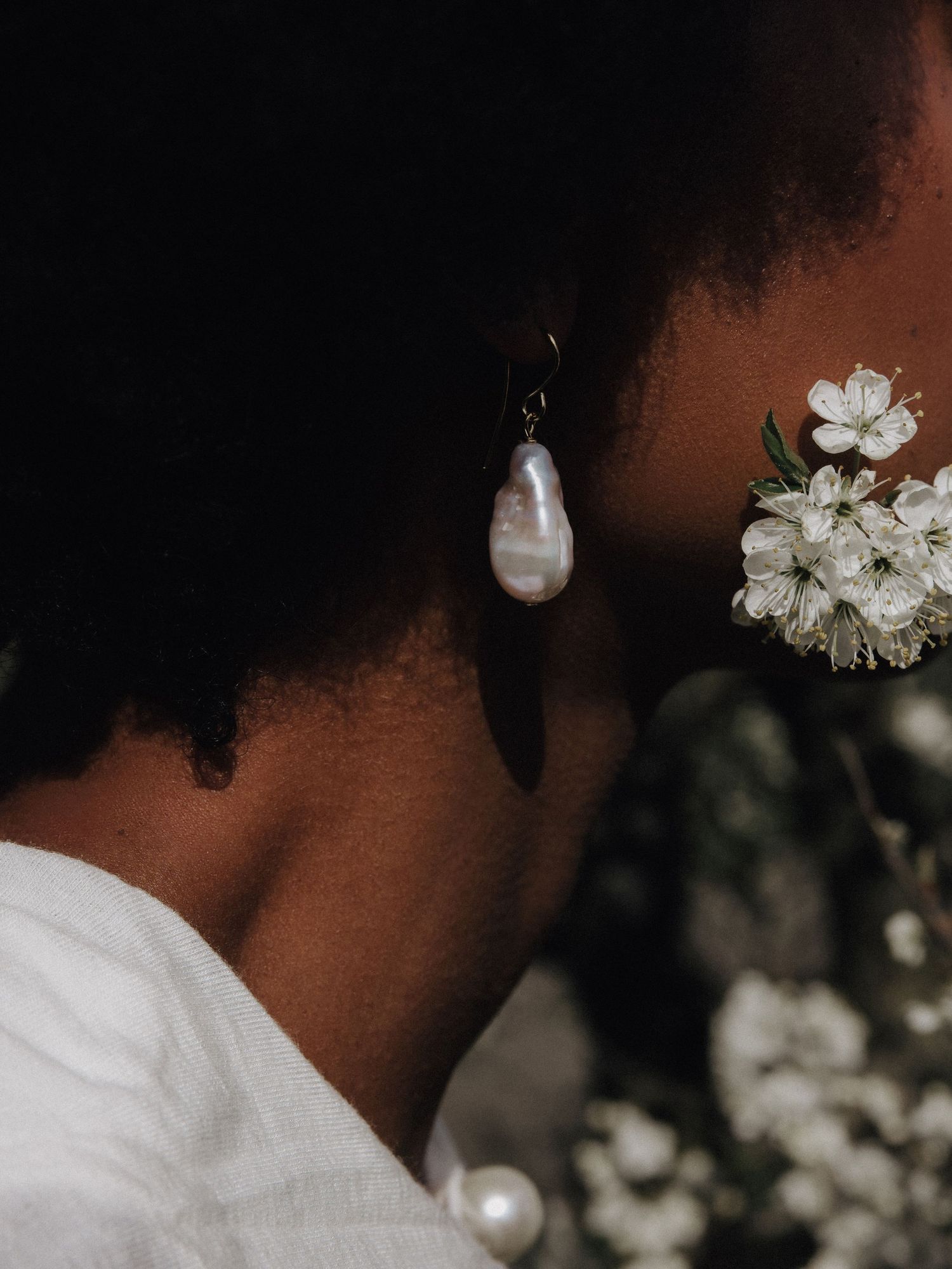 Close-up of a baroque pearl drop earring worn by a Black woman beside white cherry blossoms.