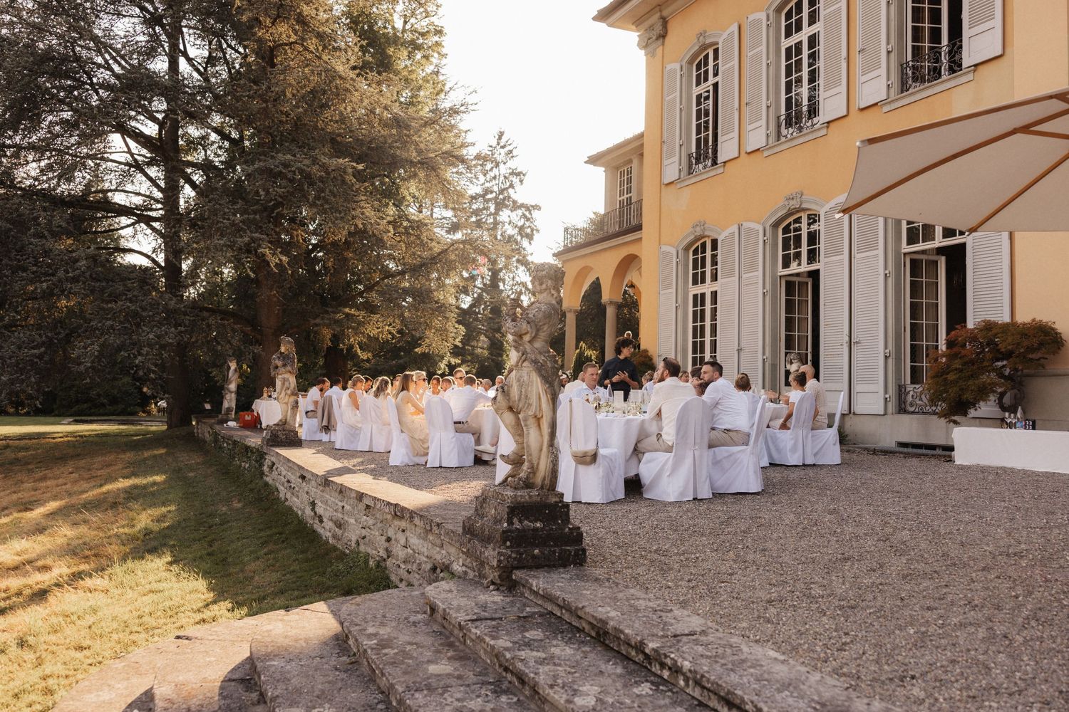 Wedding guests in white seated on a terrace of a yellow neoclassical villa surrounded by lush gardens at sunset.