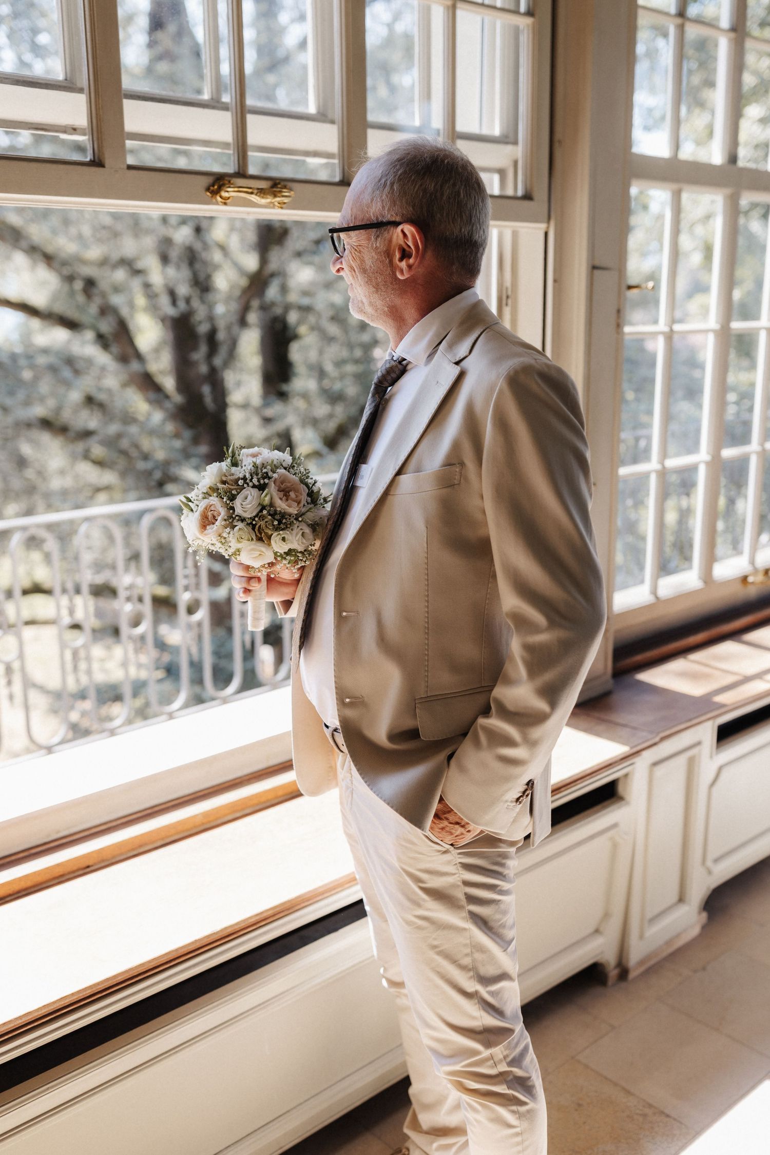 Elegant older gentleman in a beige suit holds a floral bouquet while gazing out large windows at a venue.