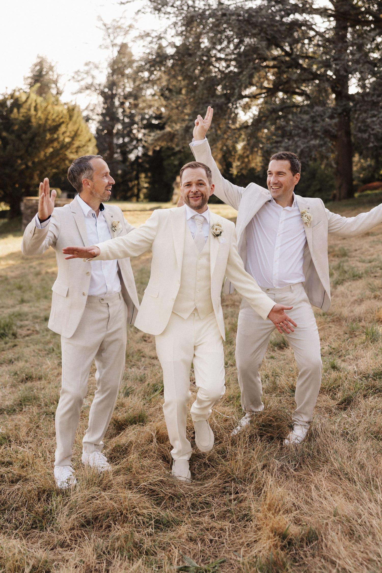 Joyful groom in cream suit jumping with two groomsmen in beige suits in a sunny outdoor setting.