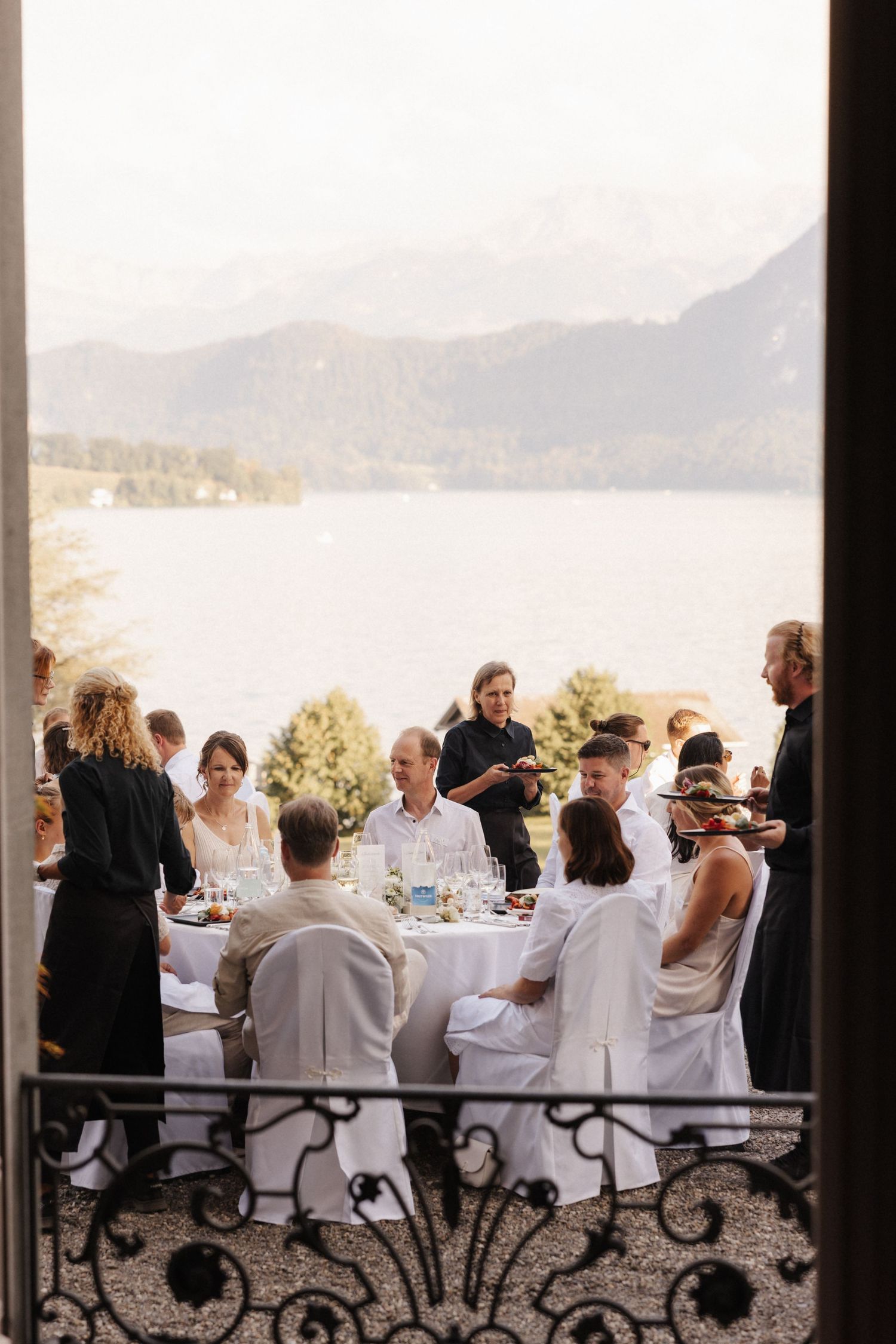 Elegant outdoor wedding reception dinner on a terrace overlooking a mountain lake with guests seated at white-clothed tables.