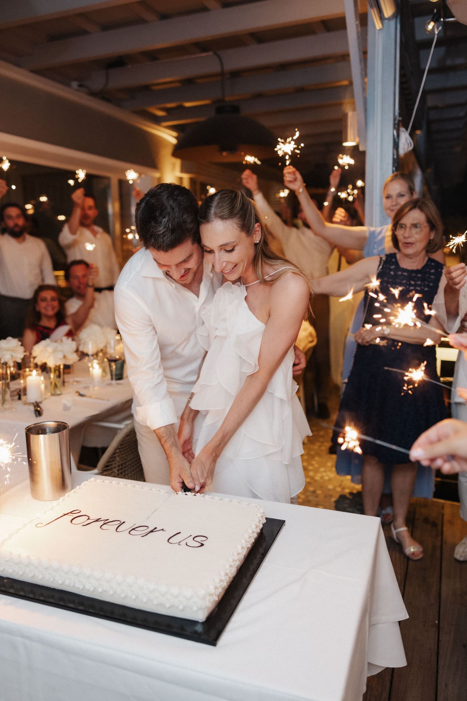 Bride and groom cutting wedding cake inscribed with forever us surrounded by sparklers.