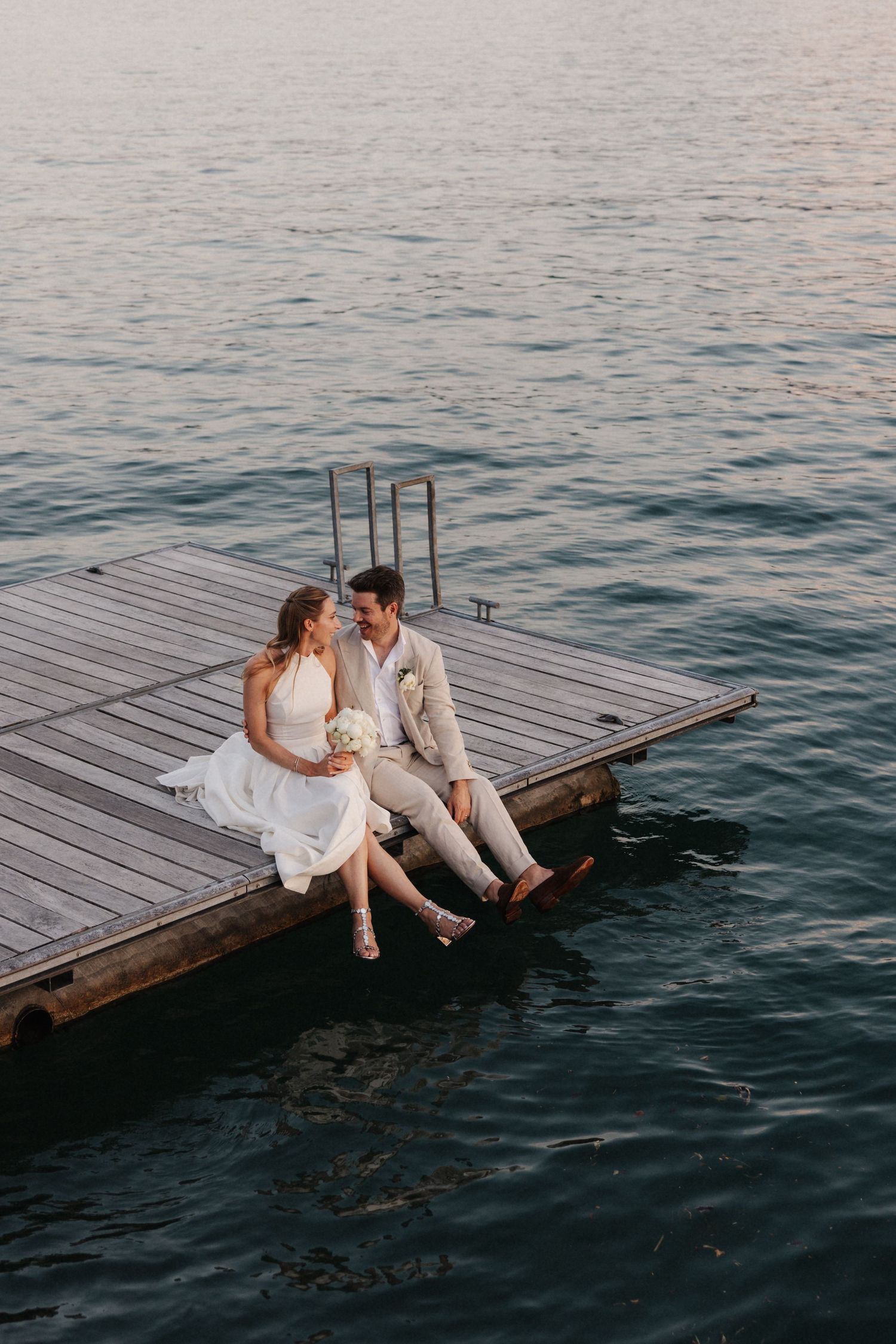 Bride and groom sit together on a wooden dock over calm water at sunset.