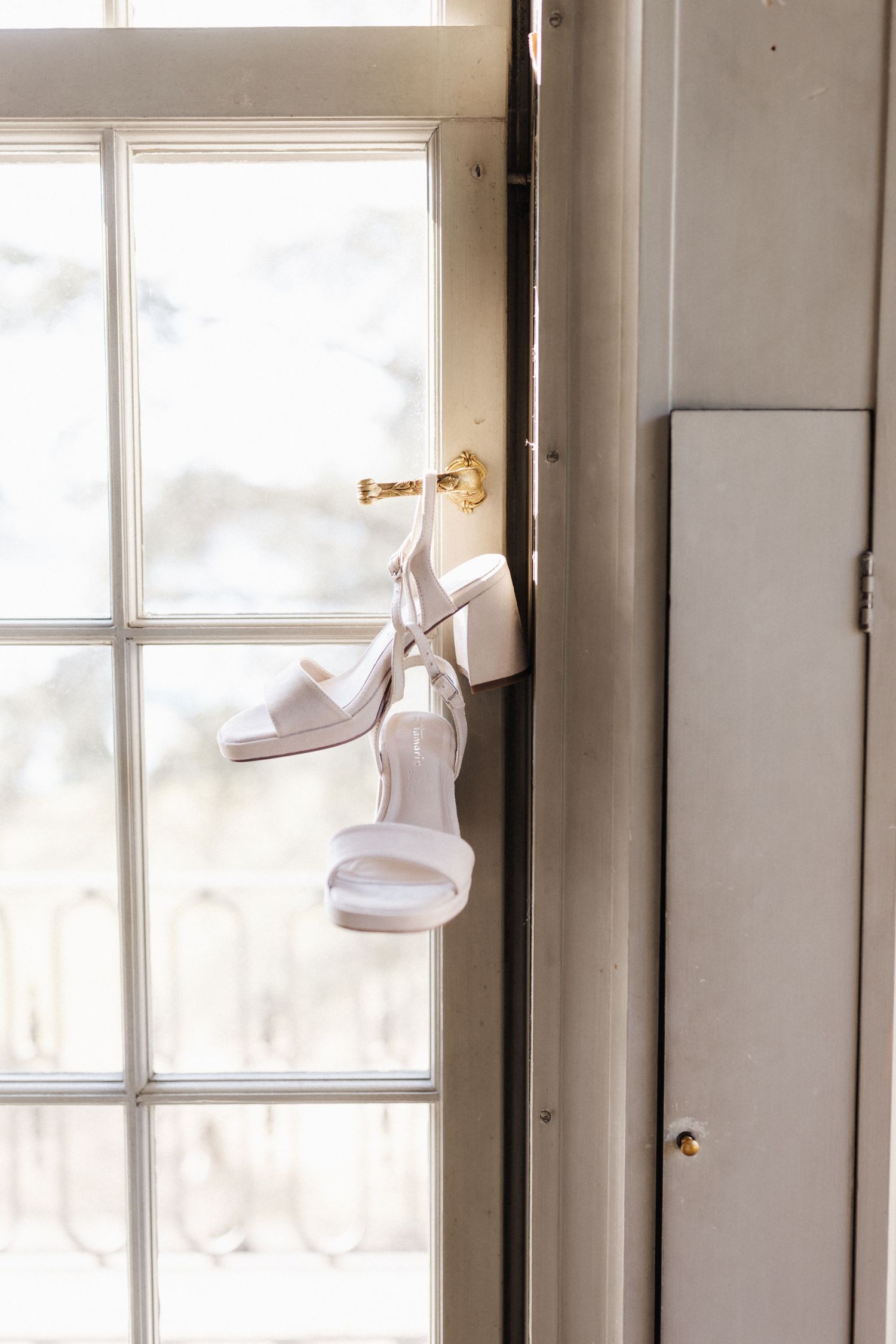 White bridal heels hanging on a gold door hook by a sunlit window.