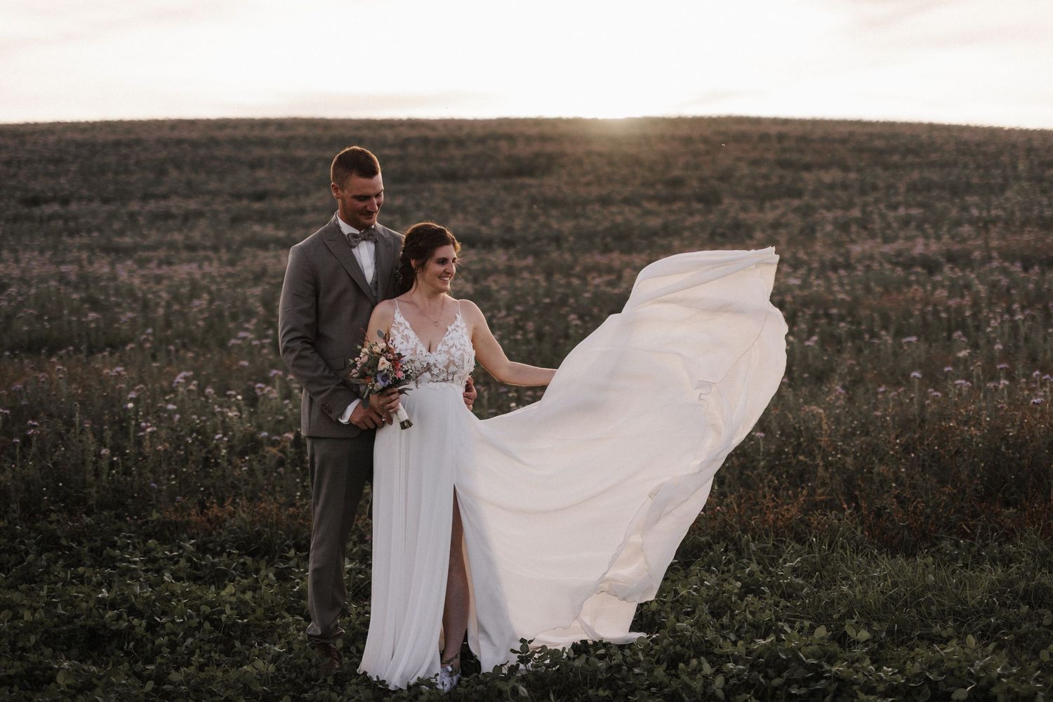 Bride twirls in flowing white gown as groom holds her close in a scenic field at golden hour.