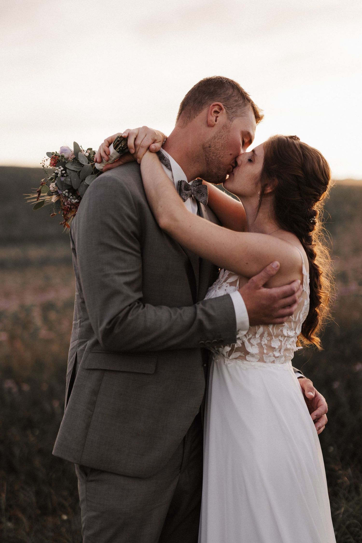 Bride and groom sharing a passionate kiss outdoors at sunset, bride holding a floral bouquet.