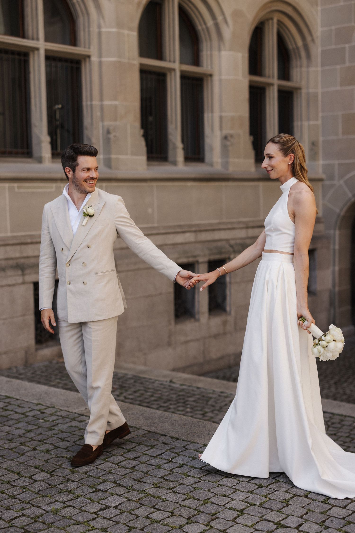 Smiling groom in beige suit holds bride's hand as she walks in white halter crop top and skirt outside historic building.