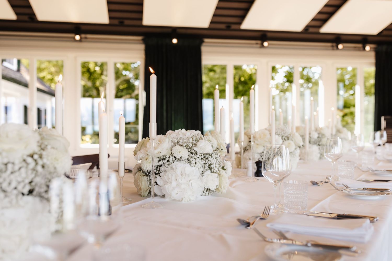 Elegant wedding reception table with white floral arrangements, tall white taper candles, and formal place settings in bright venue.