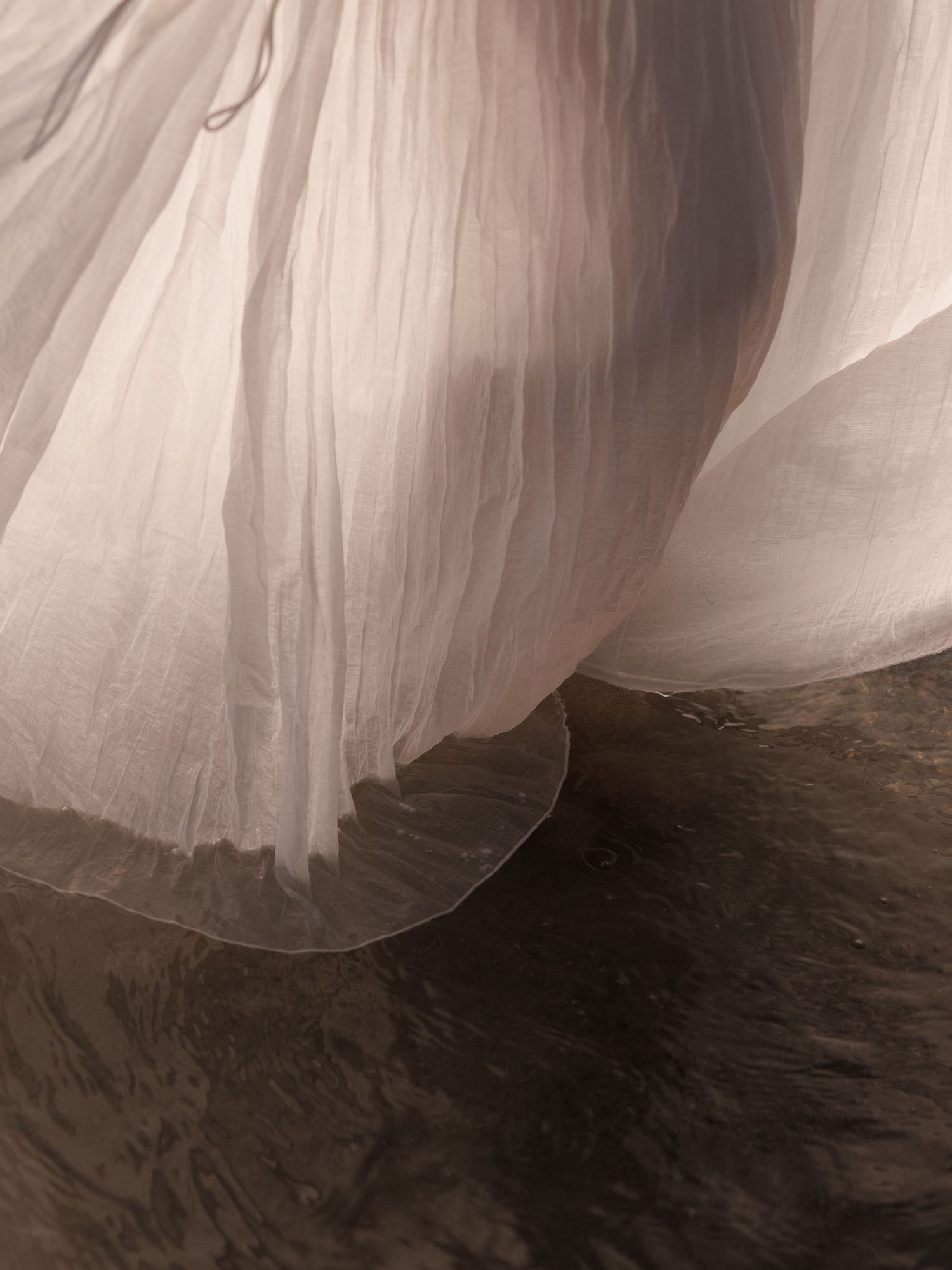 Close-up of flowing sheer white fabric skirt touching dark wet ground in soft light.