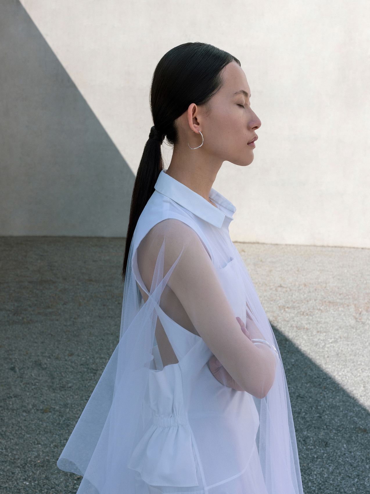 Asian woman in white sheer sleeveless top with ponytail posing in profile against concrete wall.