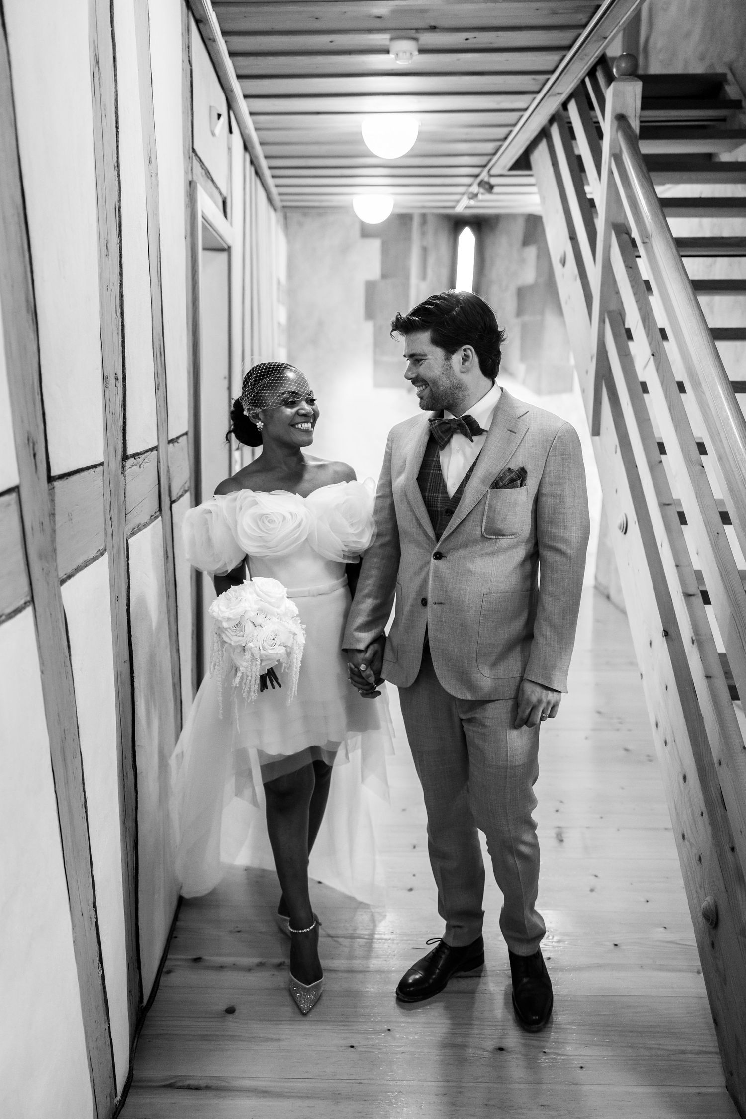 Black and white photo of happy interracial newlyweds walking hand in hand through hallway.