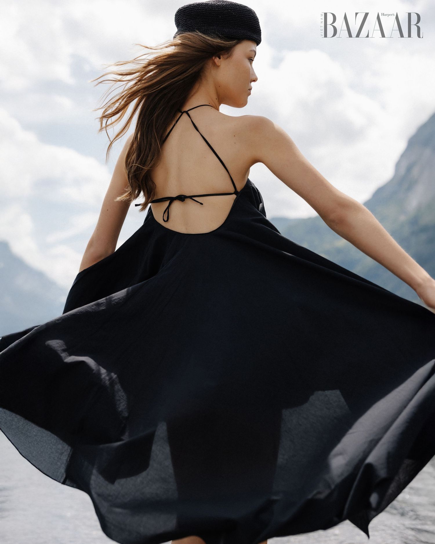 Woman in backless black dress and beret twirls with mountains and lake in background.