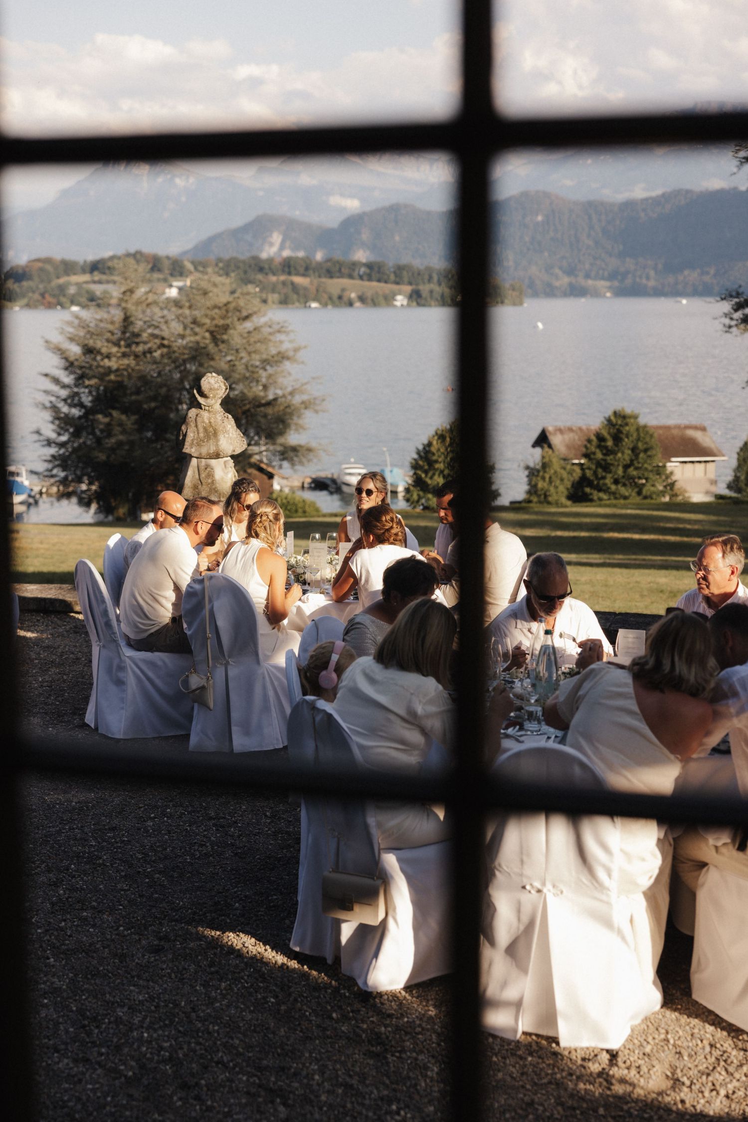 Wedding guests in white seated outdoors with a stunning alpine lake and mountain backdrop viewed through a window.