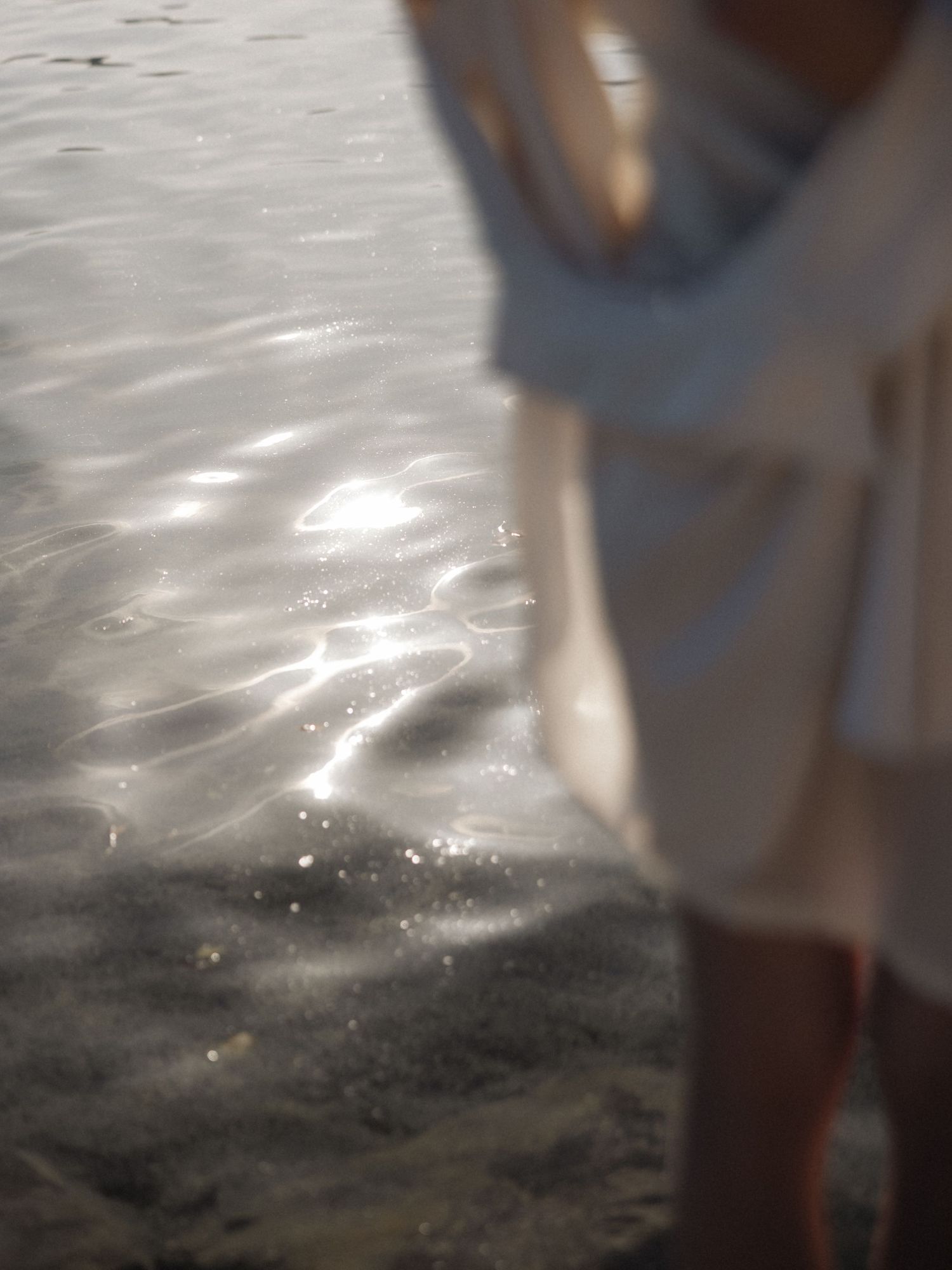 Blurred shot of person in white dress standing on sunlit sandy beach by water.
