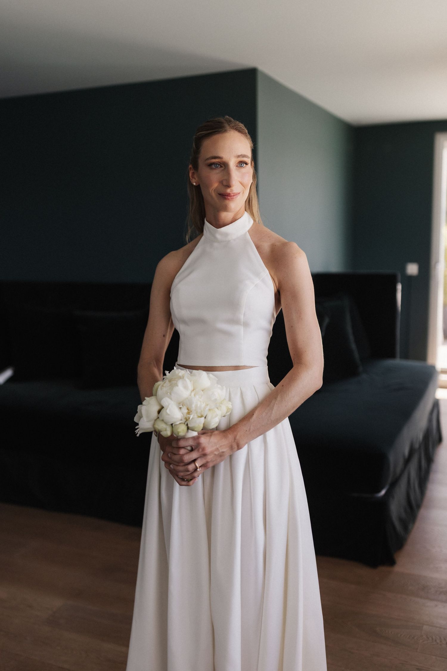 An elegant bride in a white halter-neck two-piece gown holds a small white peony bouquet indoors.