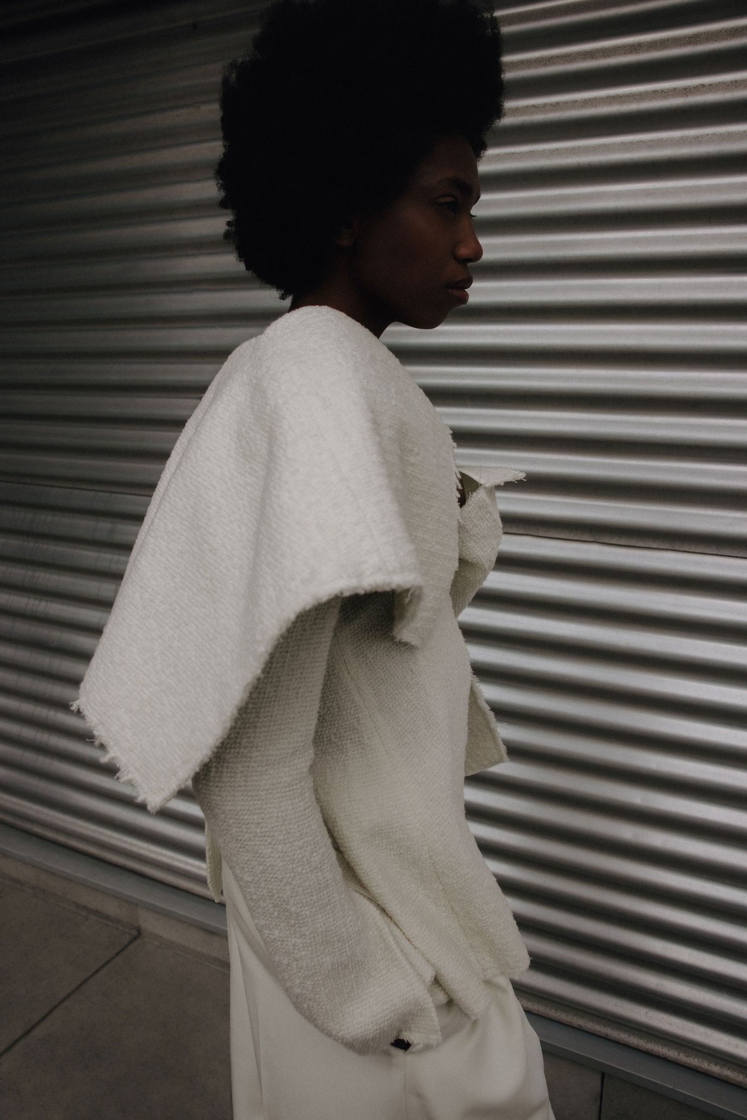 Side profile of model wearing a structured white boucle cape jacket against a corrugated metal wall.