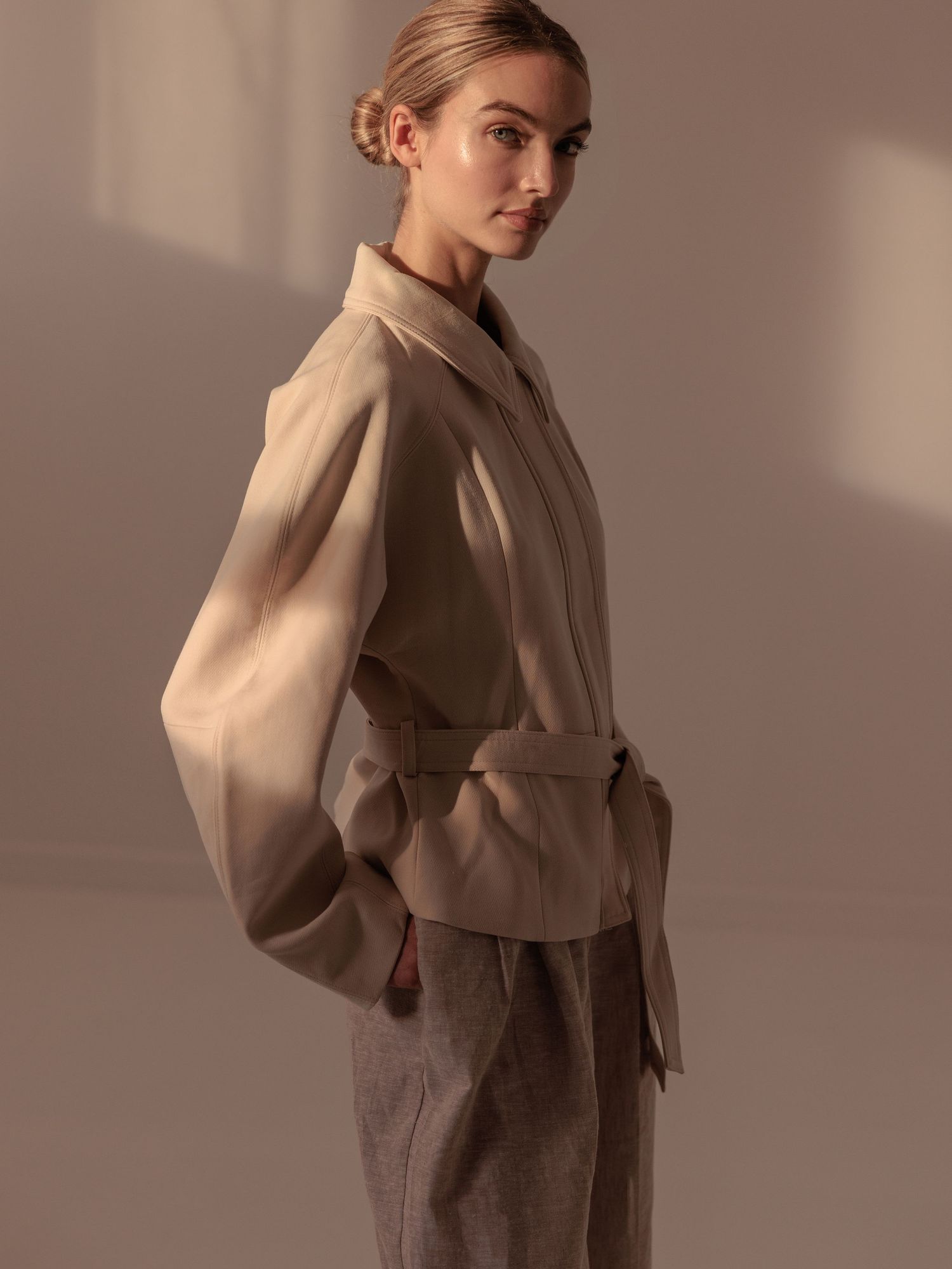 Woman in a taupe belted wrap jacket with wide sleeves stands in profile against a warm background.