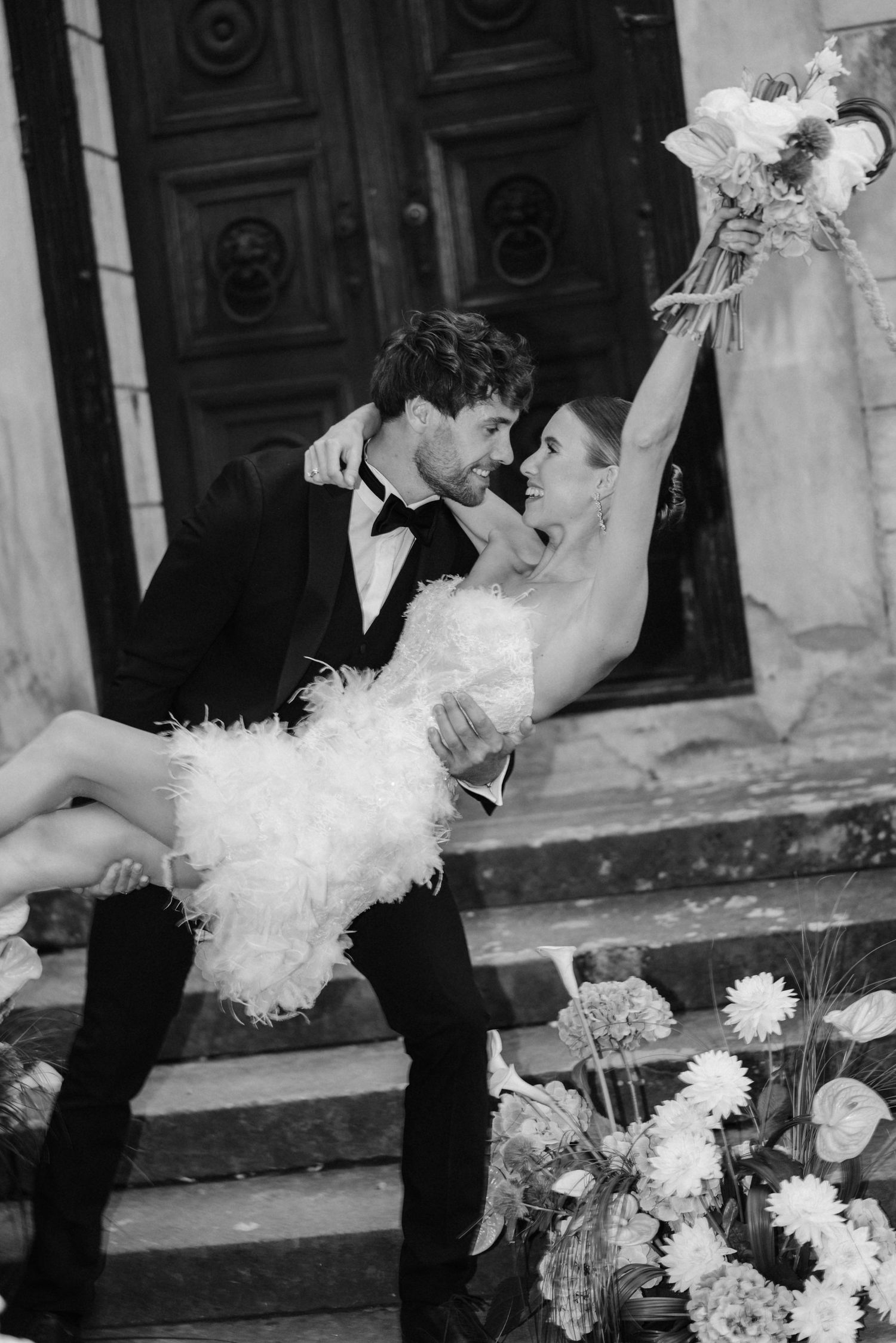 Black and white photo of groom lifting bride in feather mini dress on ornate church steps.